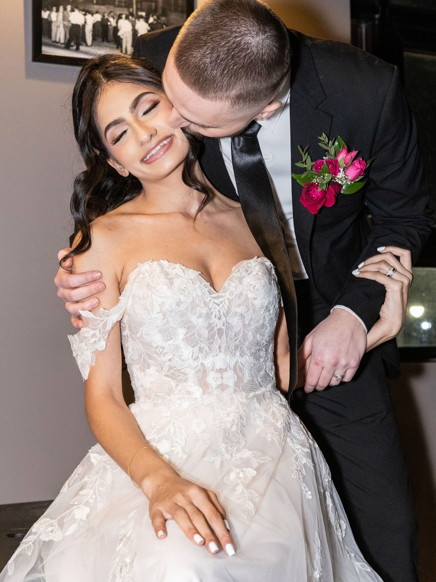 Groom in black tuxedo kissing bride in lace off-shoulder gown on the cheek at their wedding reception.