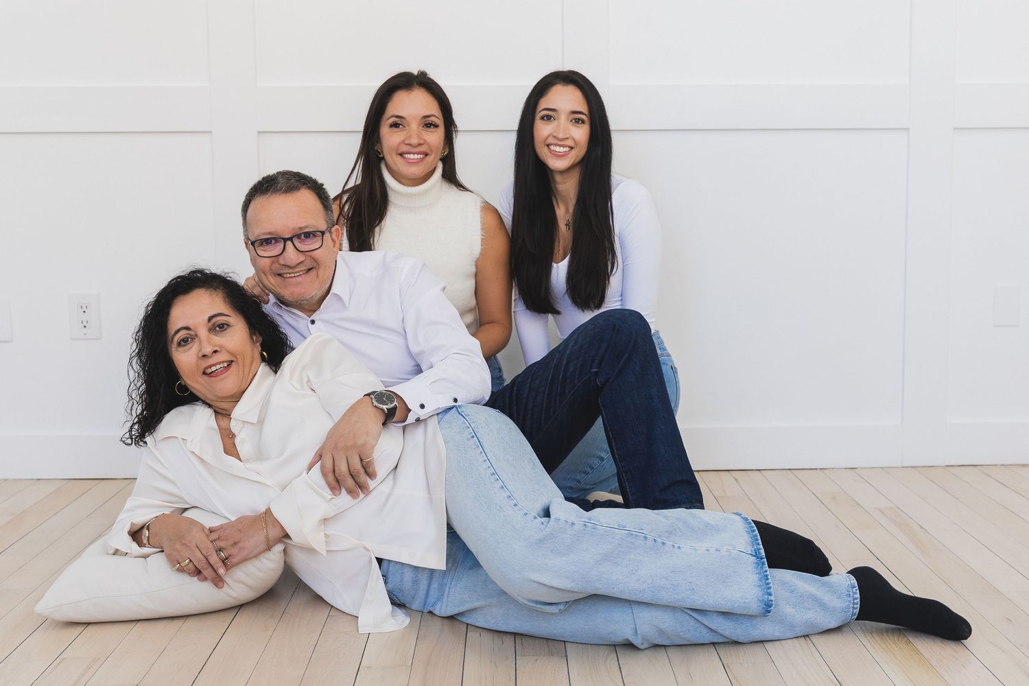 A happy family of four poses together on a light wood floor against a white wall in matching white outfits.