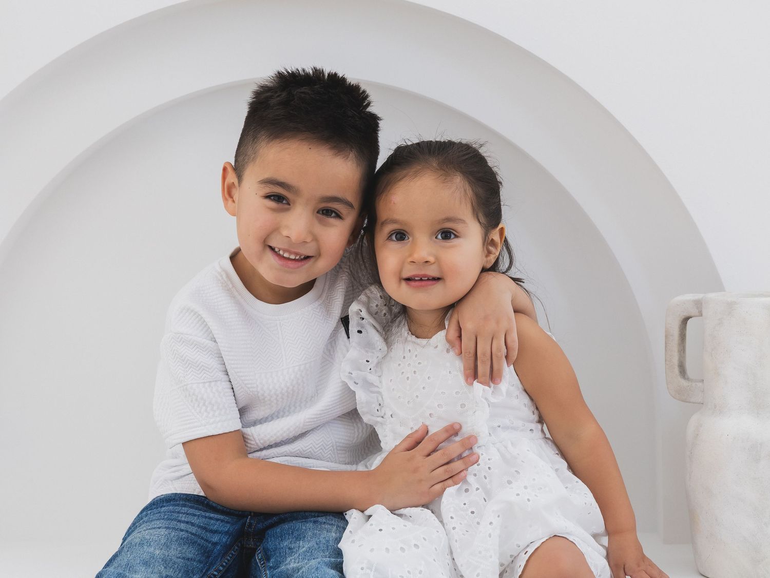 Two young siblings in white outfits pose together smiling in front of a circular white arch in a studio.