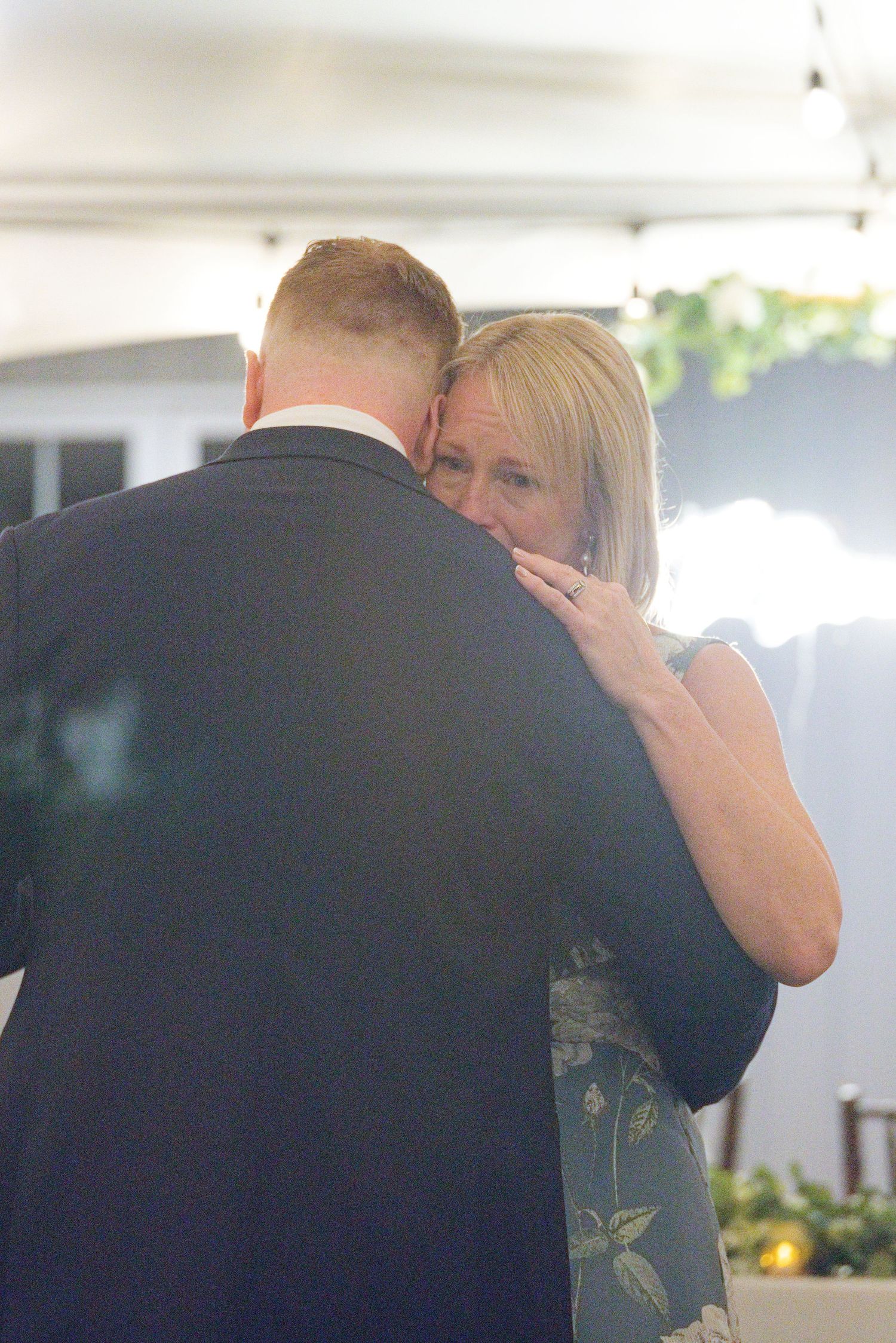 Emotional mother and son share a tender dance embrace at a wedding reception, with the mother resting her head on his shoulder.