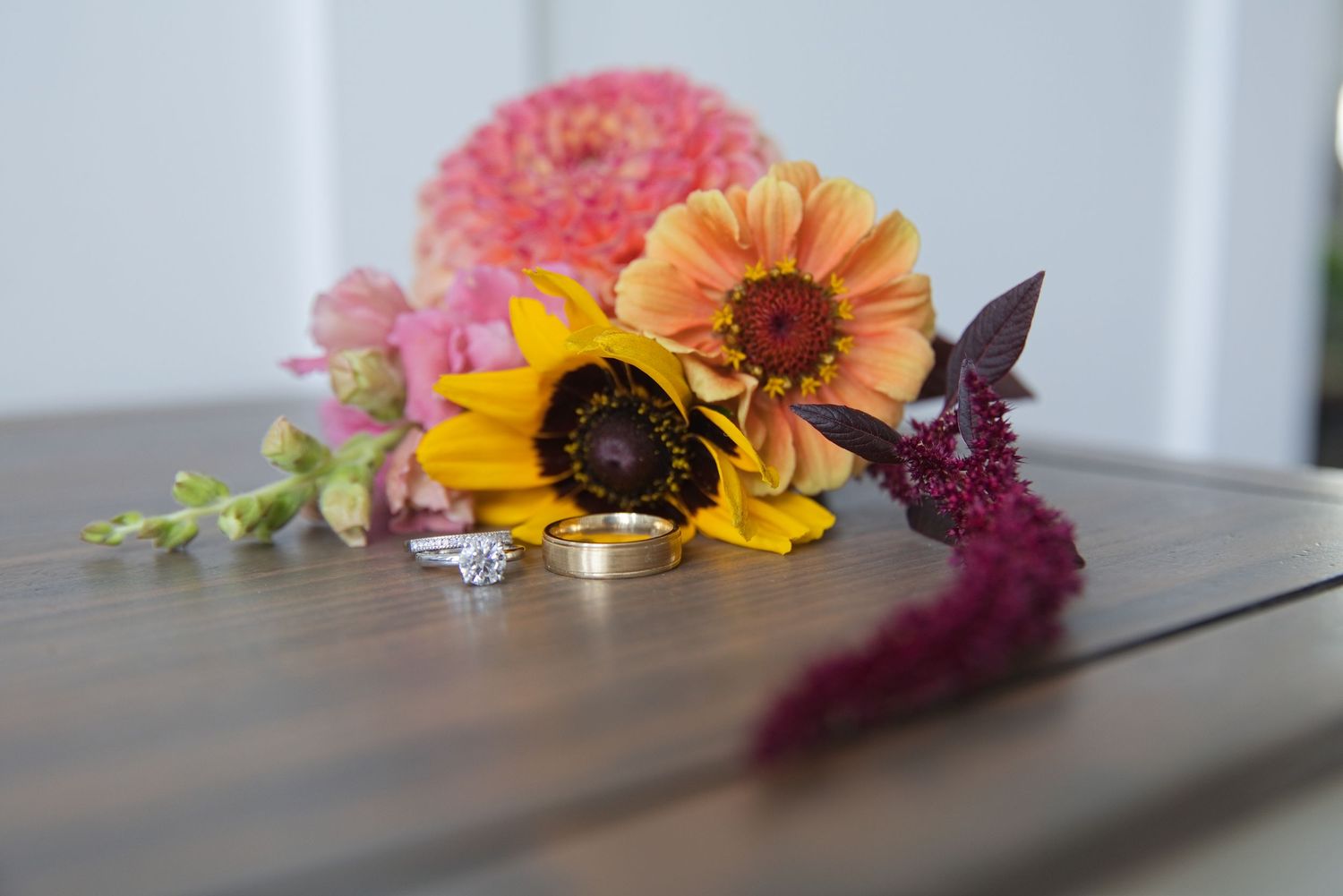 Wedding rings rest on a wooden surface surrounded by colorful fall flowers including sunflowers and pink blooms.