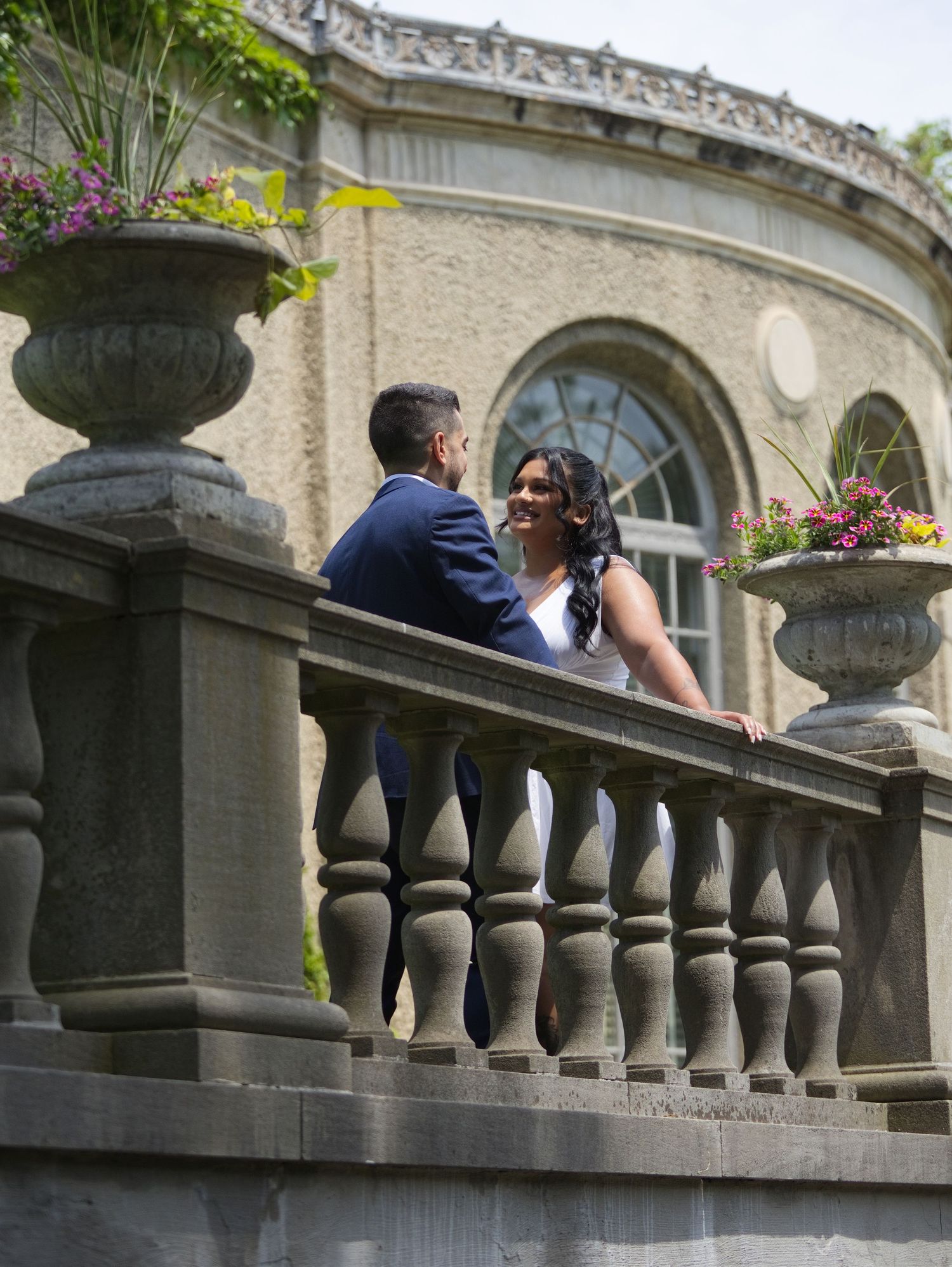 A bride and groom share a romantic moment on an elegant stone balcony adorned with floral urns at a classic venue.