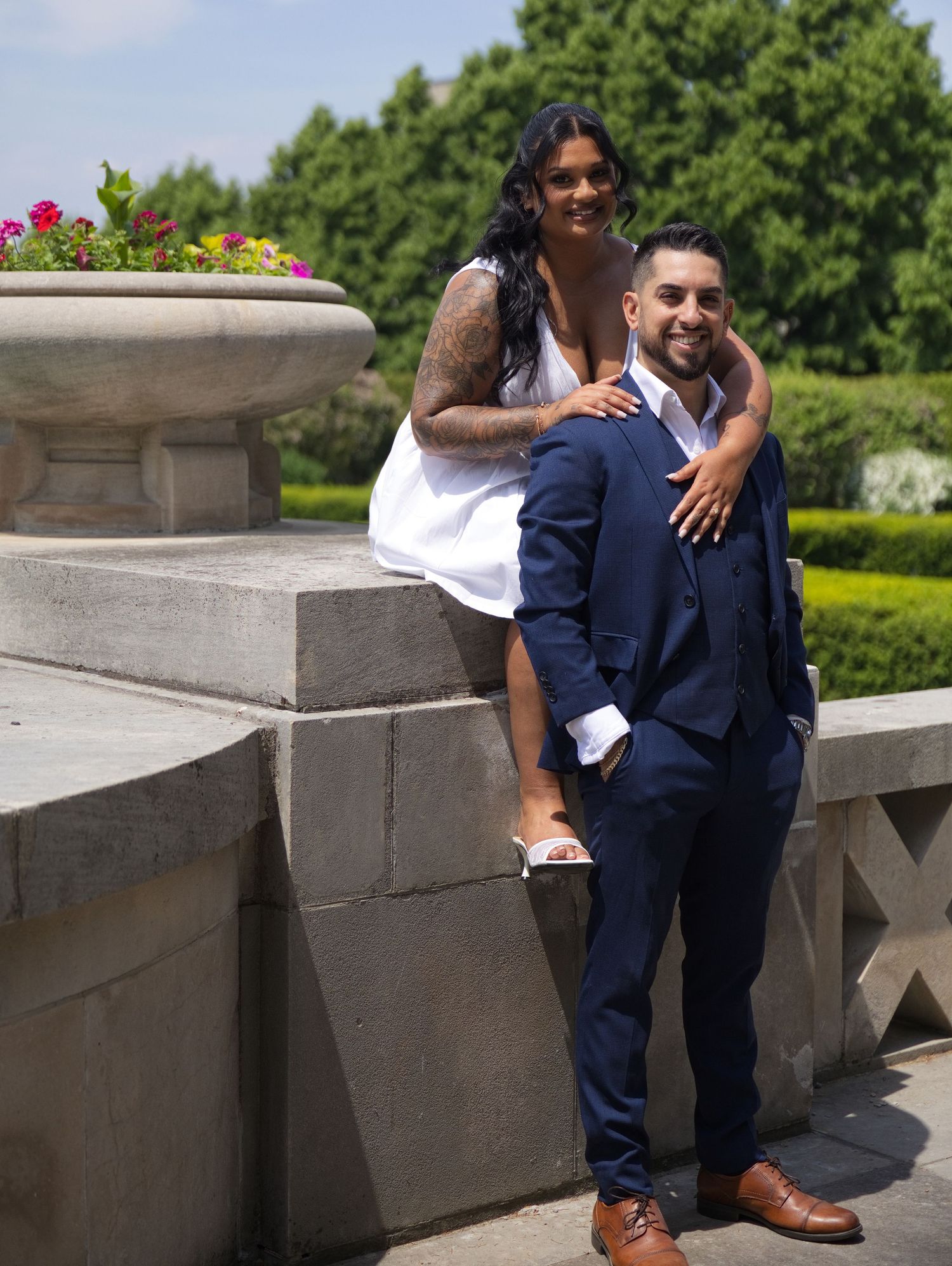 Couple posing outdoors at a garden venue, woman in white dress embracing man in navy suit on stone balustrade.
