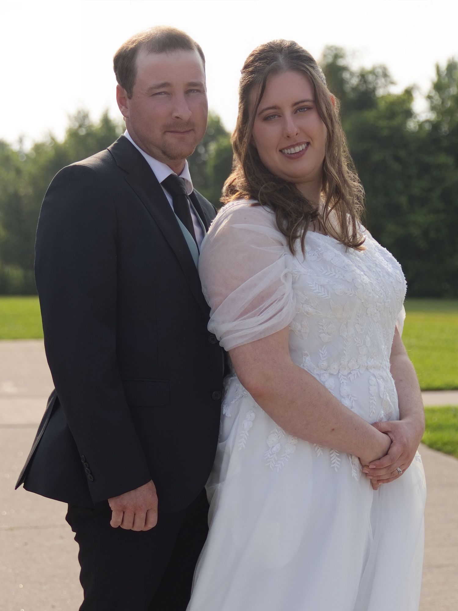 A smiling bride in a white gown and groom in a black suit posing together outdoors at their wedding.