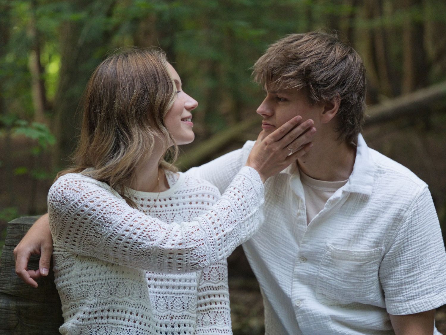 A young couple shares a tender moment outdoors, with a woman gently touching a man's face near a wooden fence.