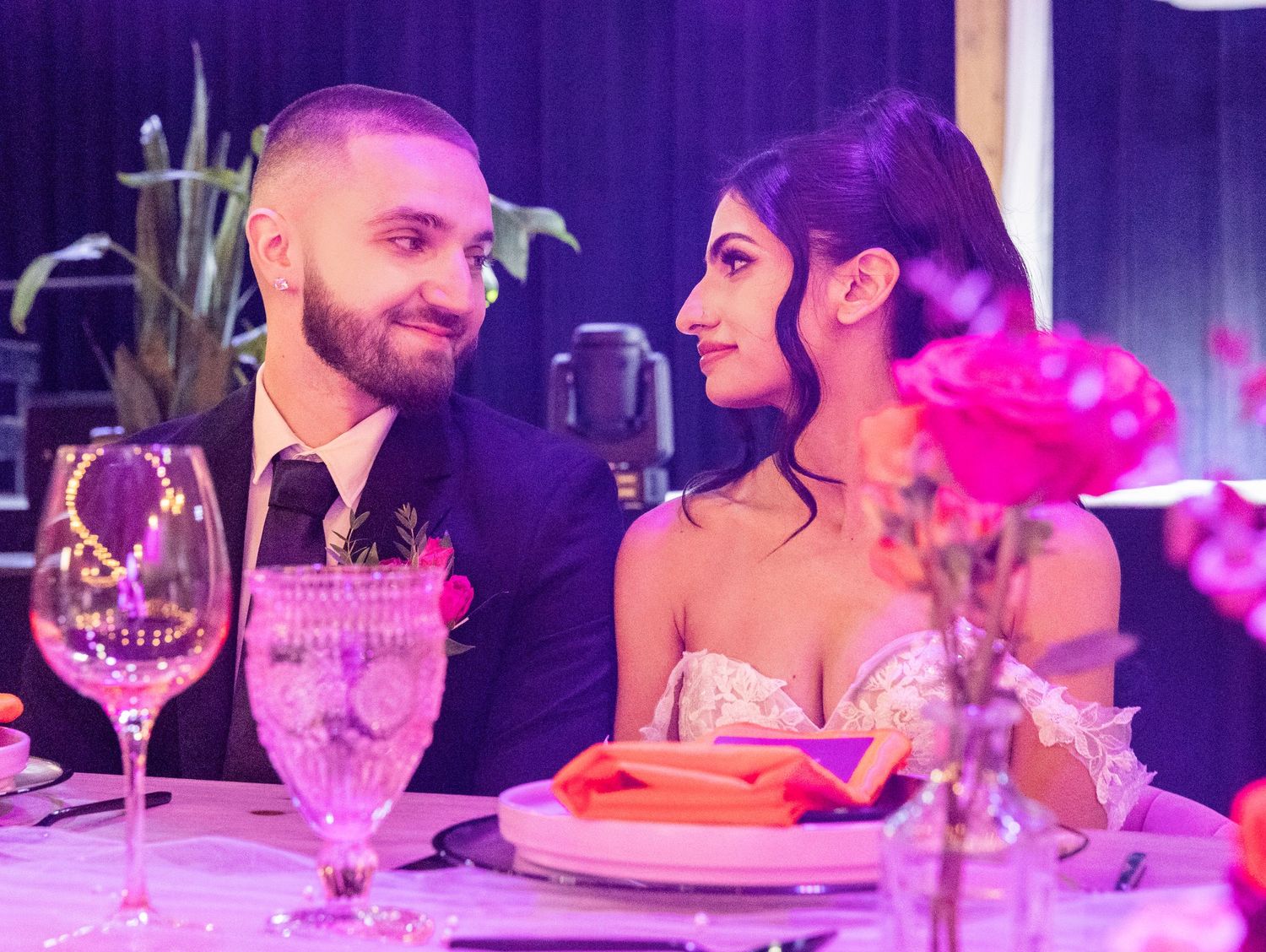 Bride and groom share a romantic glance at sweetheart table adorned with pink floral arrangements.