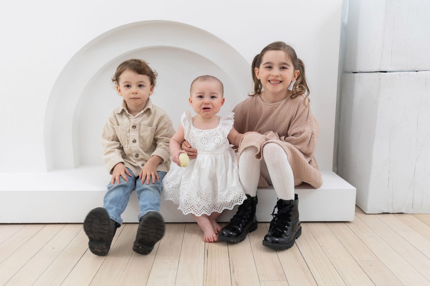Three young children, including a baby in a white dress, sit together on a white bench in a bright studio.