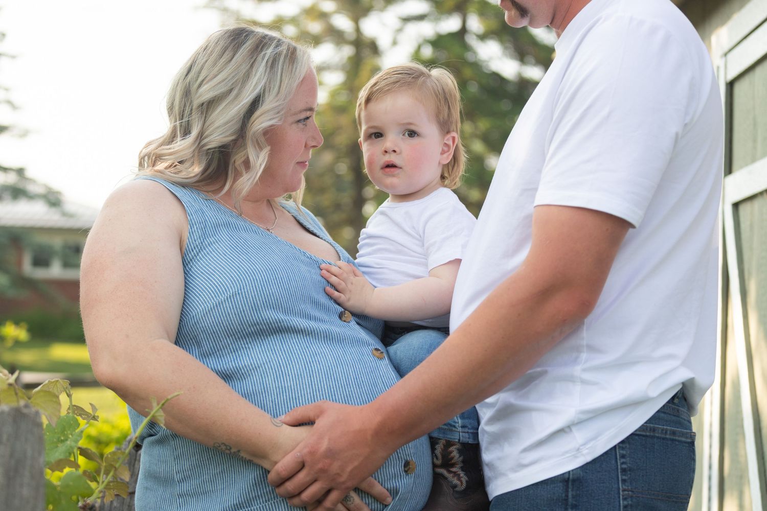 A pregnant woman in a blue dress holds a toddler alongside a man in a white t-shirt outdoors.