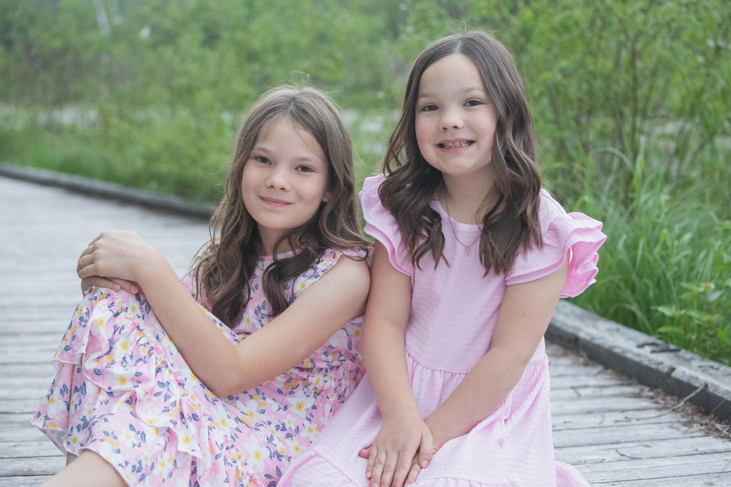 Two young girls in pink dresses smile together on a stone path surrounded by greenery.
