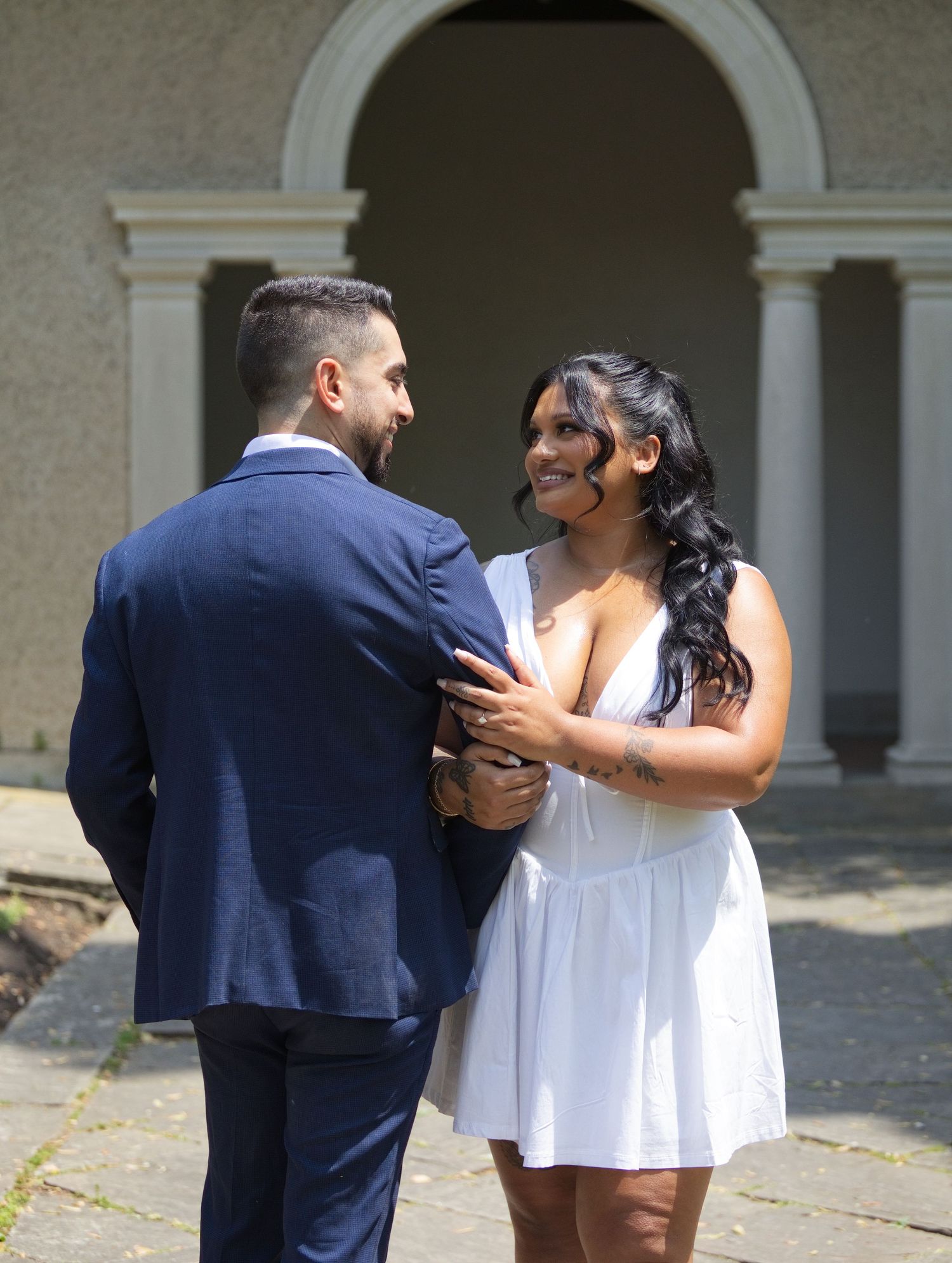 Smiling couple in white dress and navy suit embracing before arched stone building.