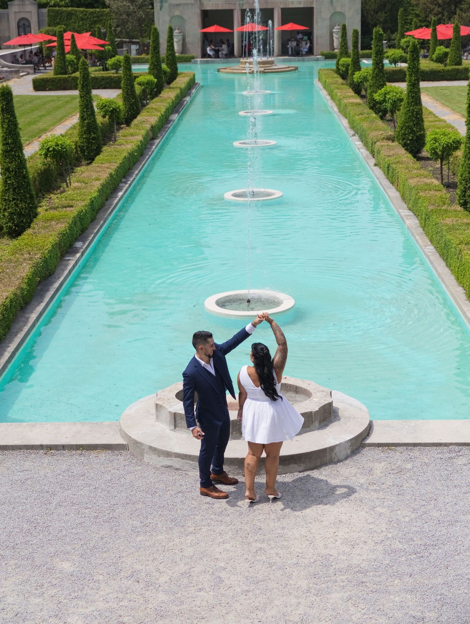 Couple dancing by a turquoise fountain pool in a formal garden with manicured hedges and red umbrellas.