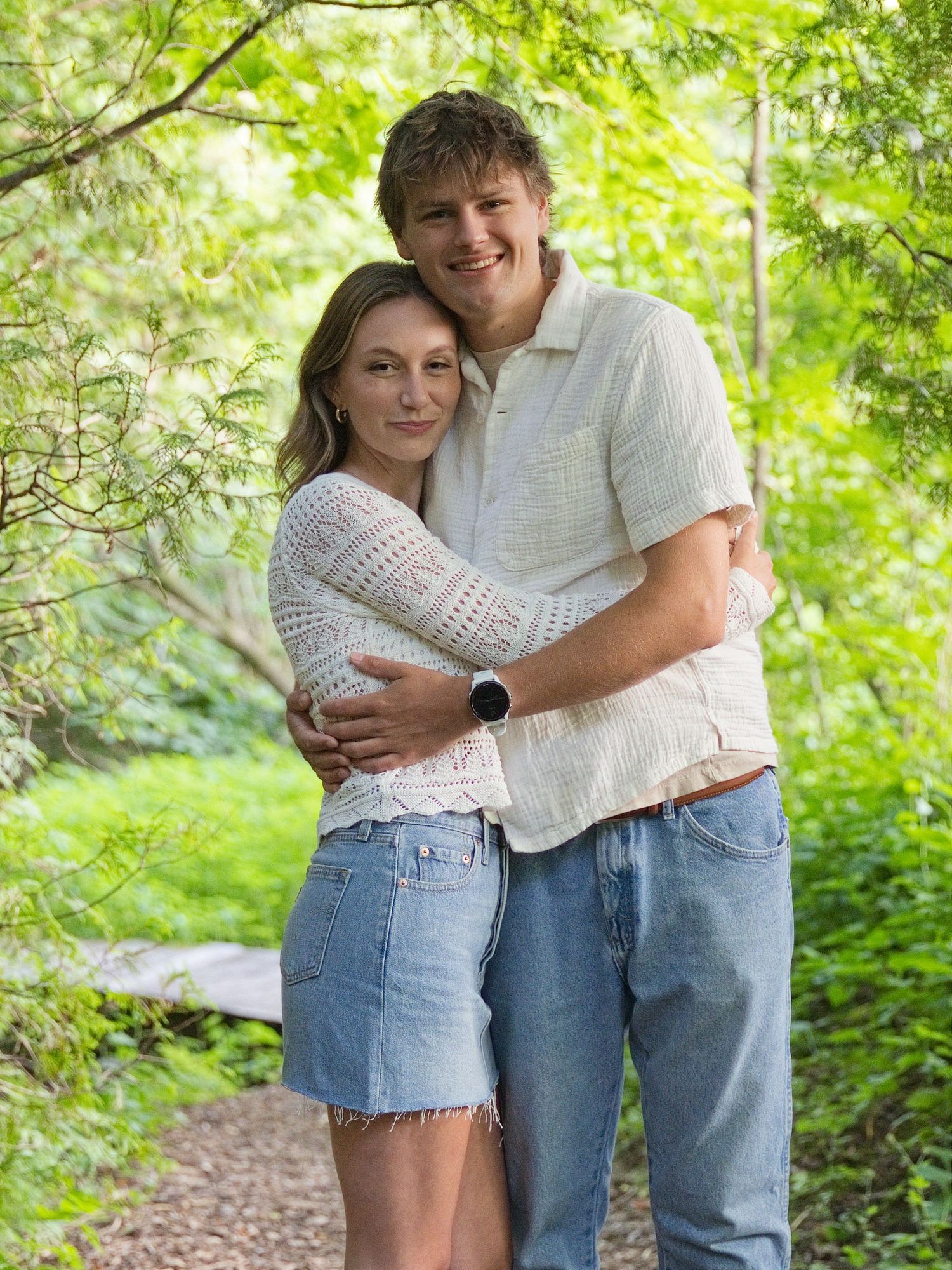 A young couple embraces and smiles on a wooded garden path, wearing casual denim and light tops.