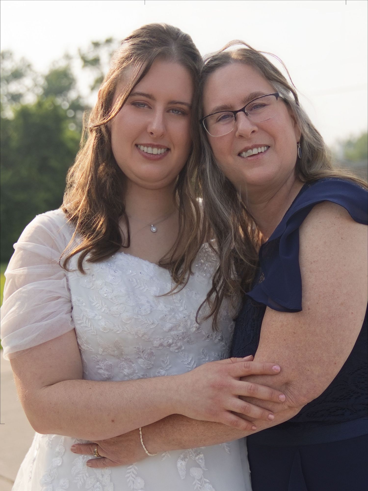 Bride in white lace gown smiles with older woman in navy dress outdoors at wedding celebration.