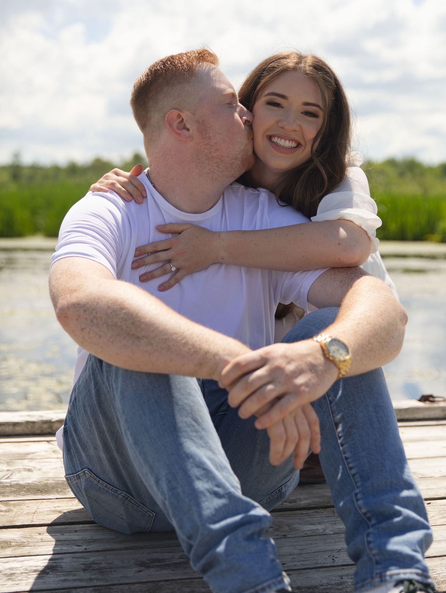 A couple sits on a wooden dock by a lake, sharing a kiss, wearing white shirts and jeans on a sunny day.