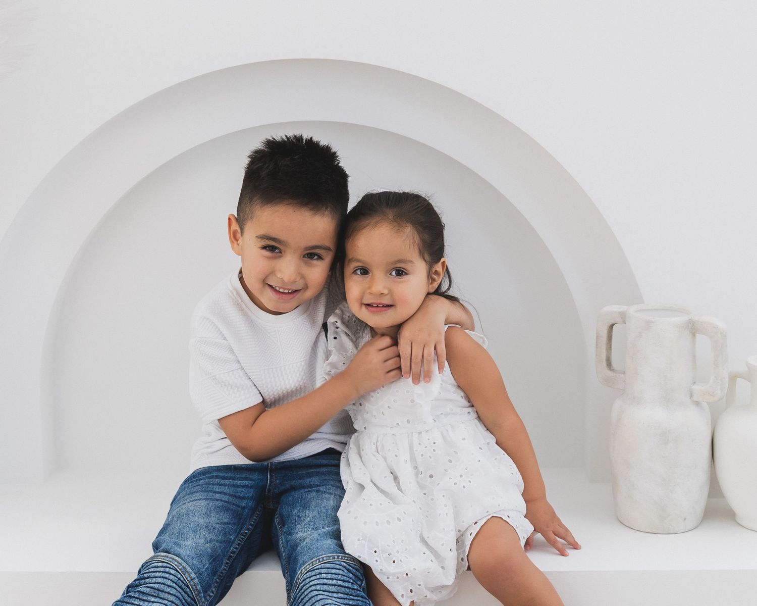 A cheerful boy in a white shirt embraces his younger sister in a white dress against a white arch backdrop.
