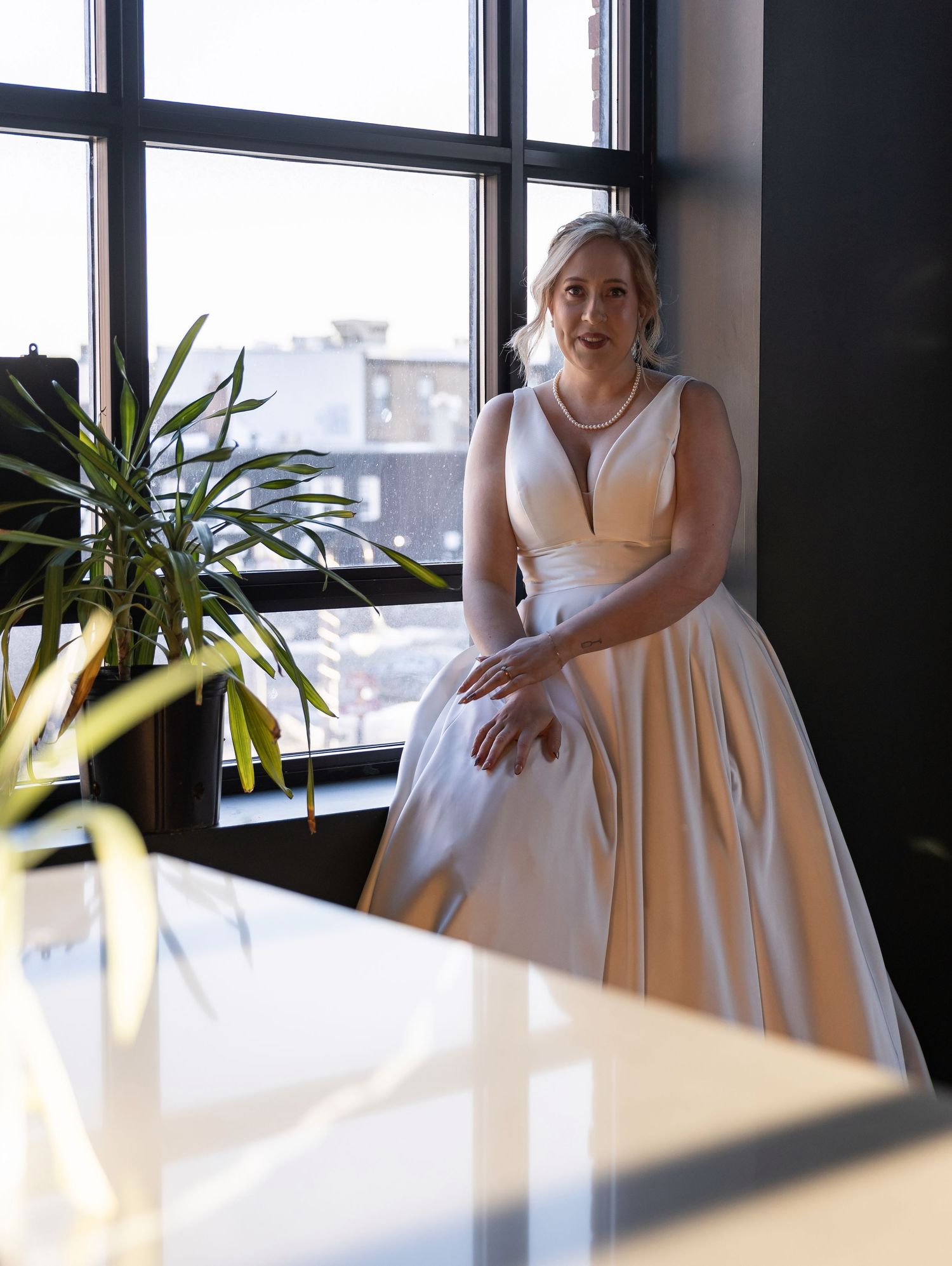 Elegant bride in a champagne satin ball gown standing by large windows with natural light streaming in.