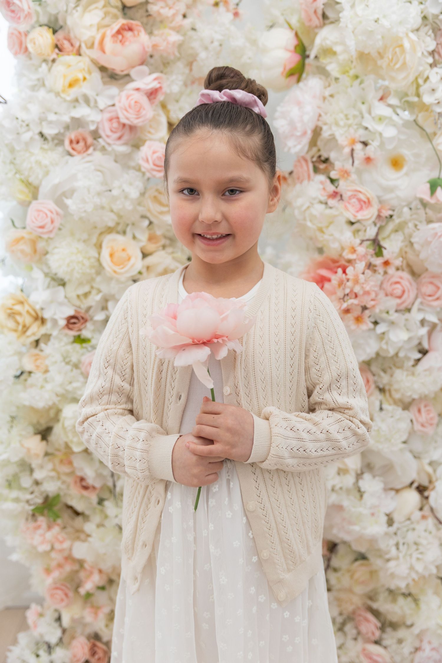 Young girl in white dress holding a pink flower in front of floral wall backdrop.