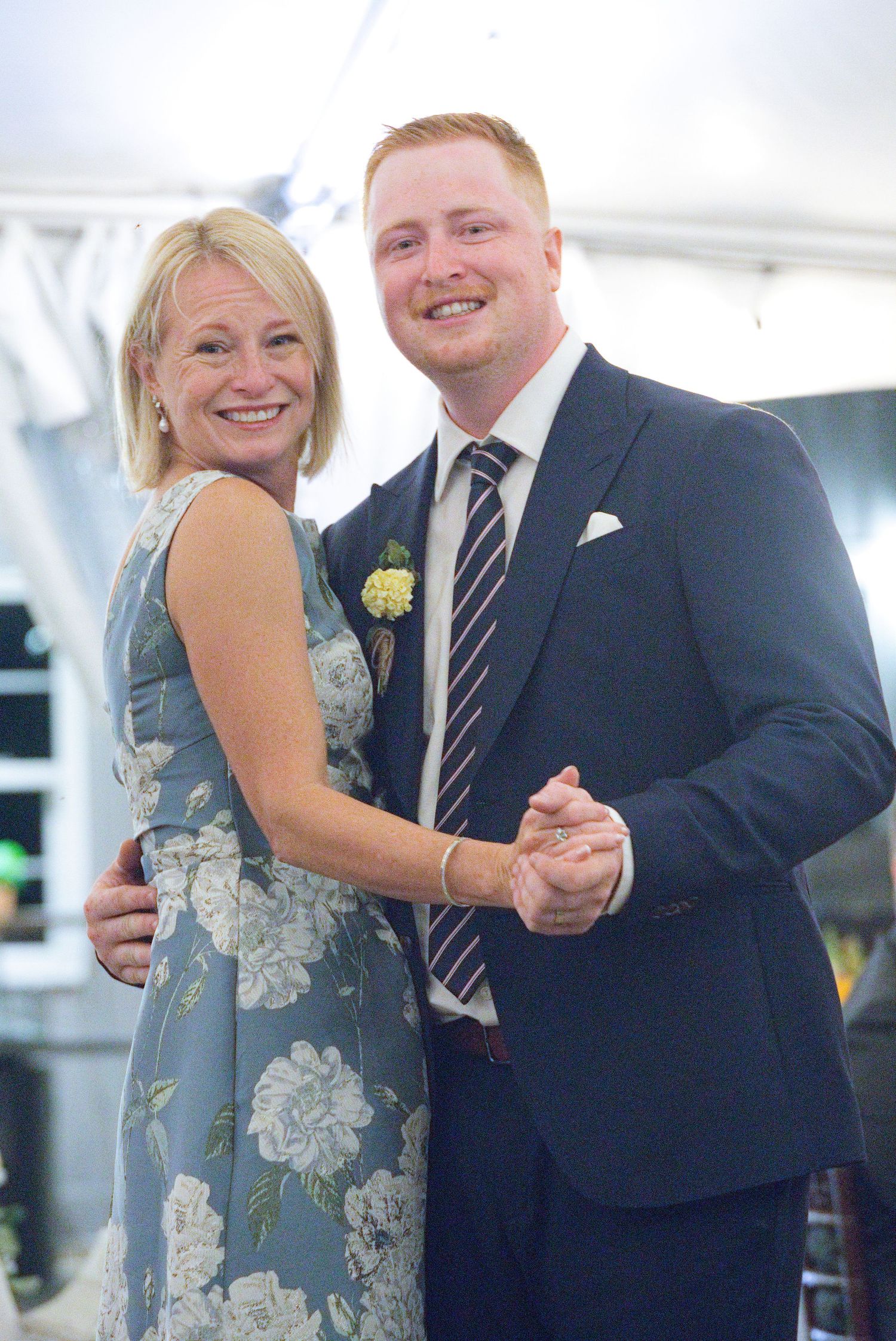 A smiling couple dancing at a wedding reception, man in navy suit with boutonniere, woman in floral blue dress.