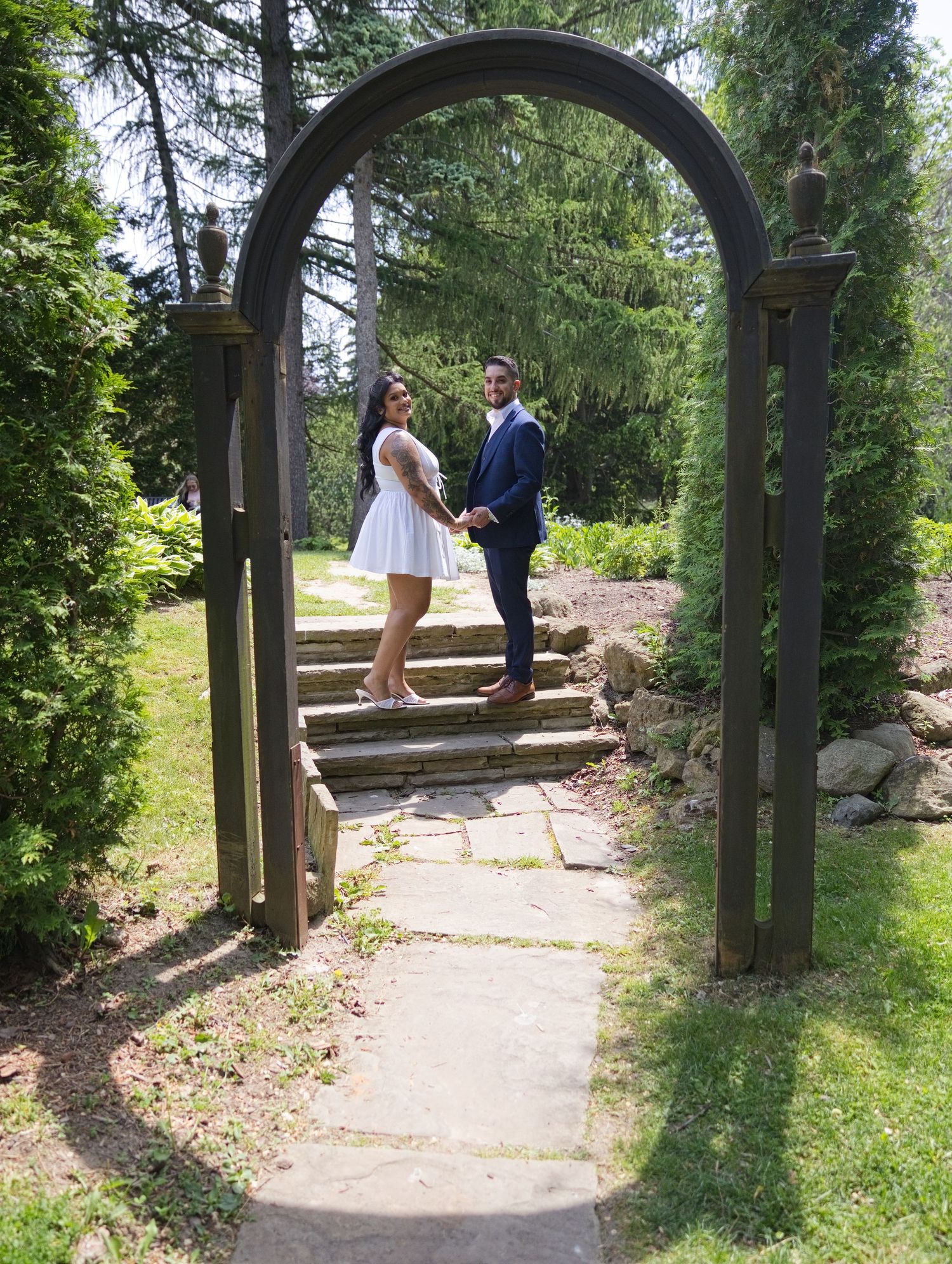 Couple in wedding attire posing under a wooden garden arch on stone steps surrounded by lush greenery.