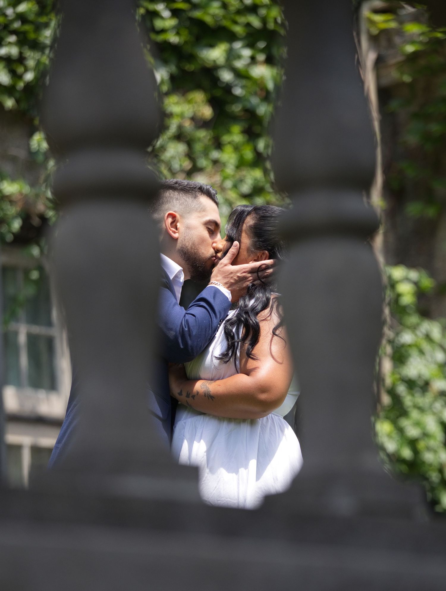 Couple sharing a romantic kiss framed by decorative iron railings with lush green foliage in the background.