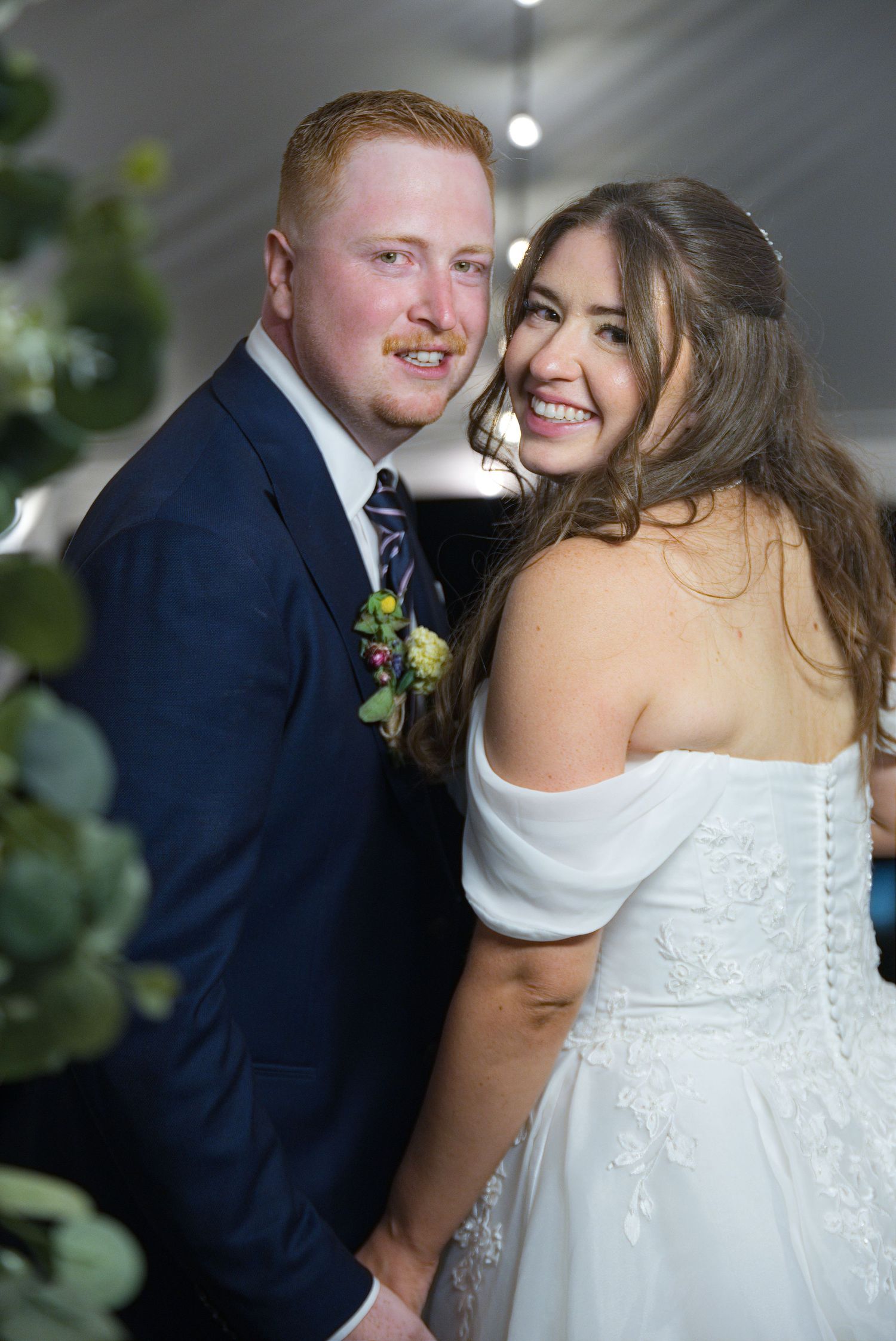 Smiling bride in white off-shoulder gown and groom in navy suit with boutonniere pose together at wedding reception.
