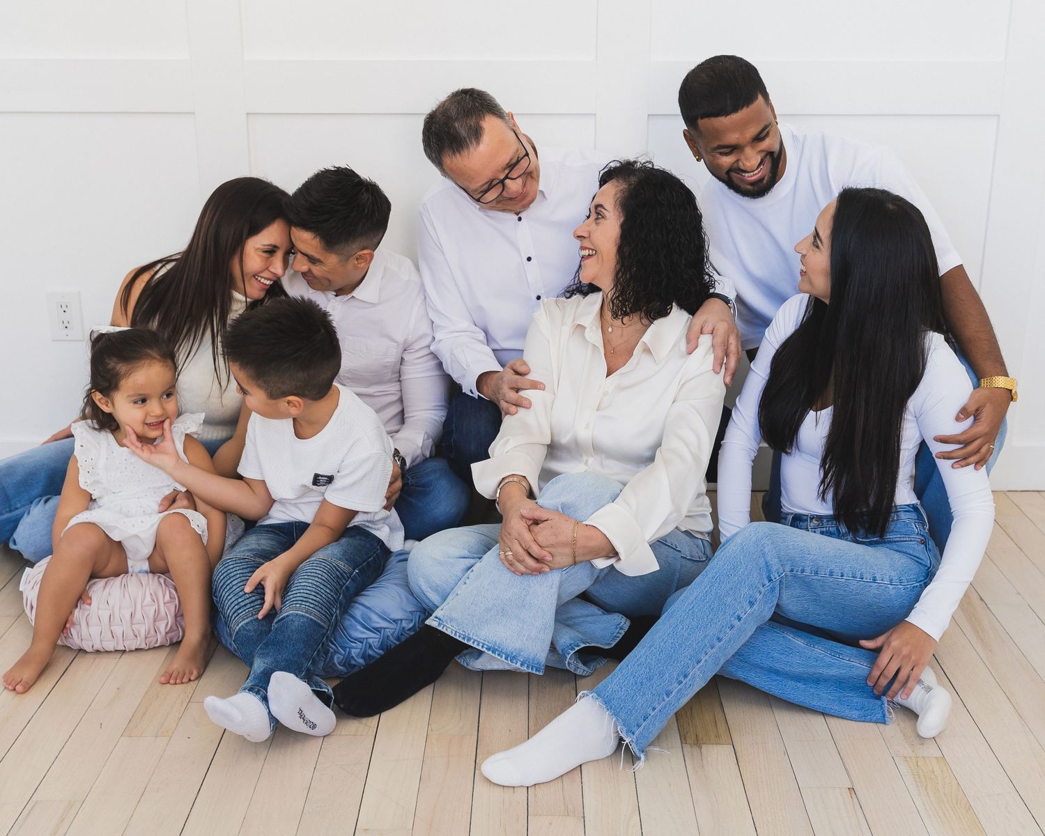 A joyful multigenerational family laughing together, wearing white tops and jeans, sitting on a light wood floor.