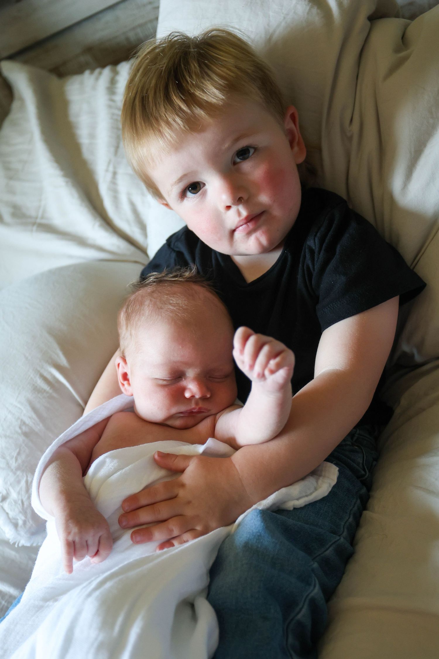 Young boy holding sleeping newborn baby sibling on bed.