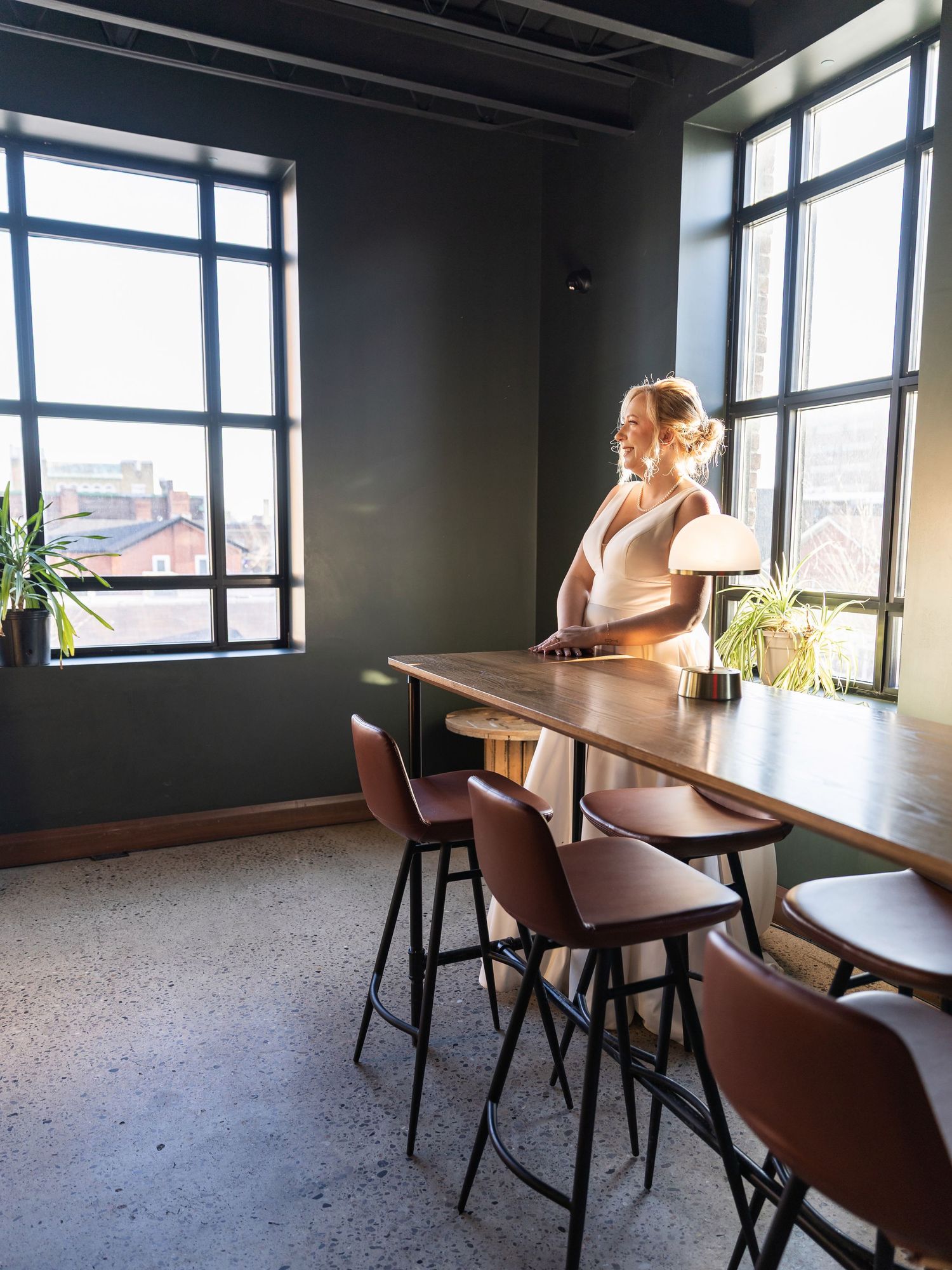 Bride in white dress standing by wooden bar table near large industrial windows with city view.