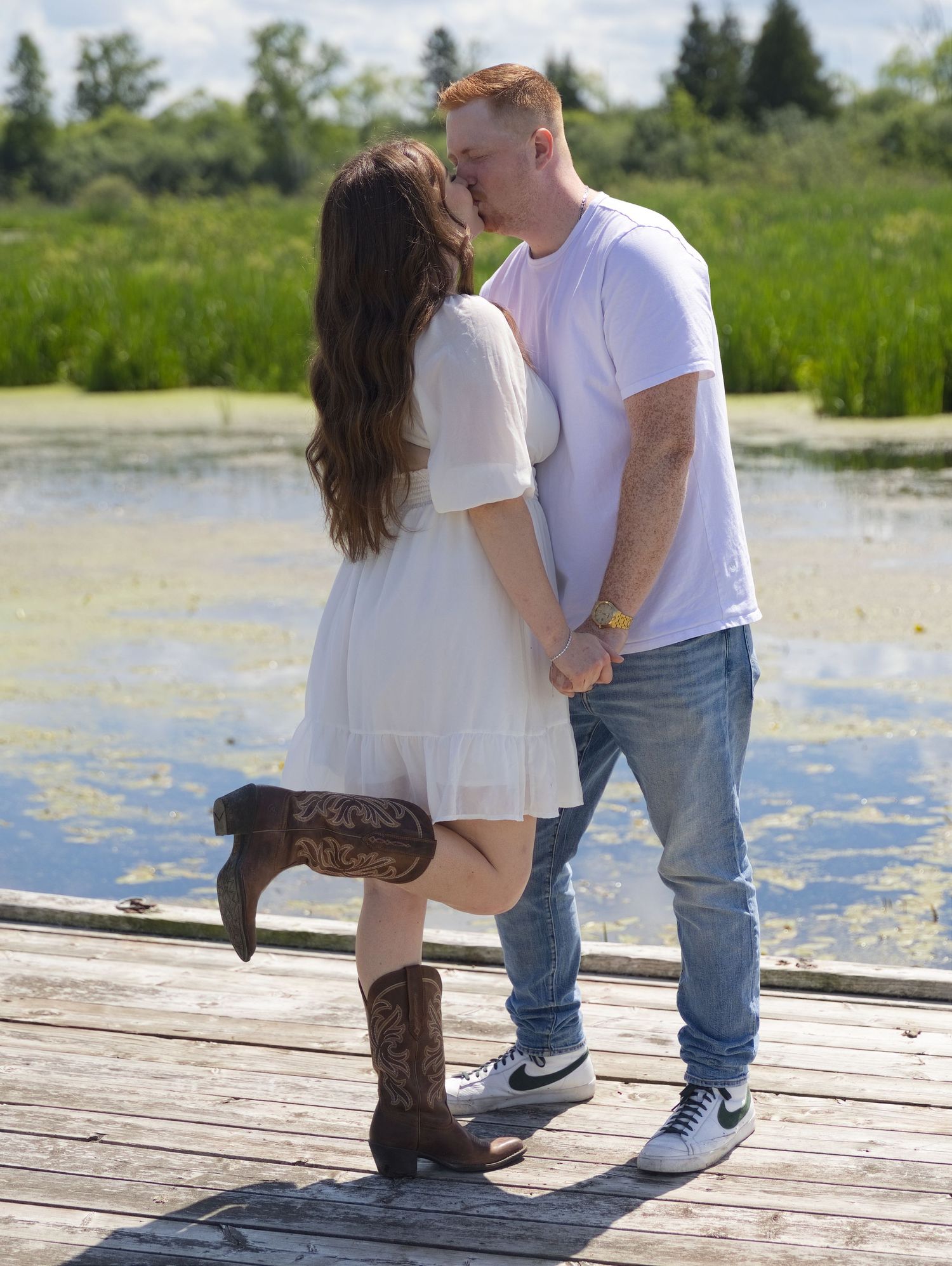 Couple kissing on a wooden dock by a pond, woman in white dress and cowboy boots, man in white tee and jeans.