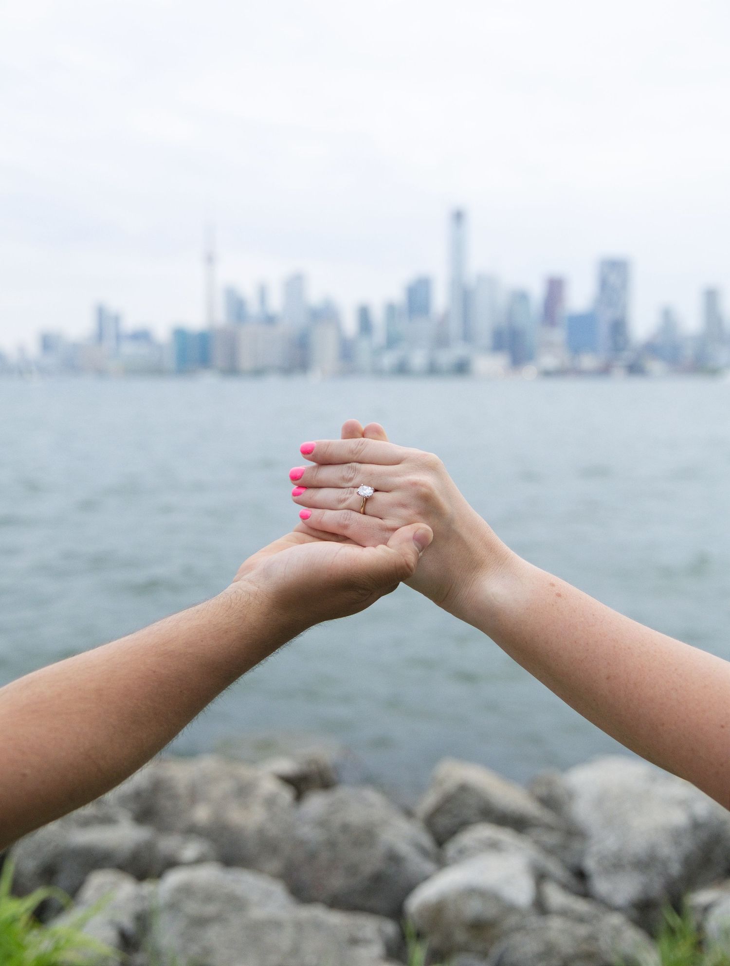 Two hands clasped together showing an engagement ring with a city skyline and waterfront in the background.