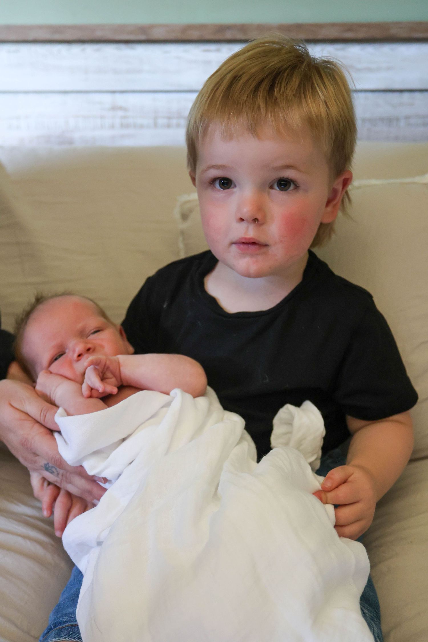 A young toddler boy in a black shirt gently holds a swaddled newborn baby while sitting on a couch.