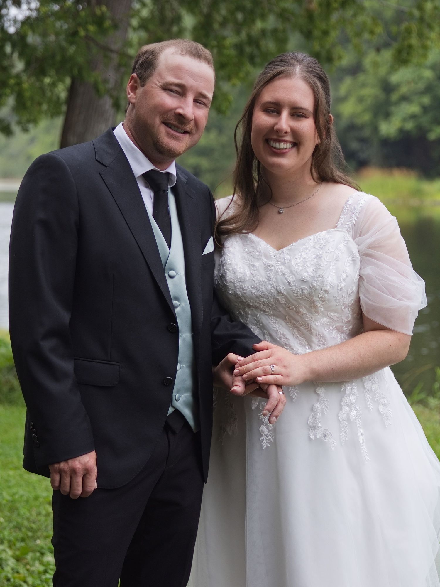 Smiling bride and groom posing together outdoors in wedding attire, white lace gown and black tuxedo.
