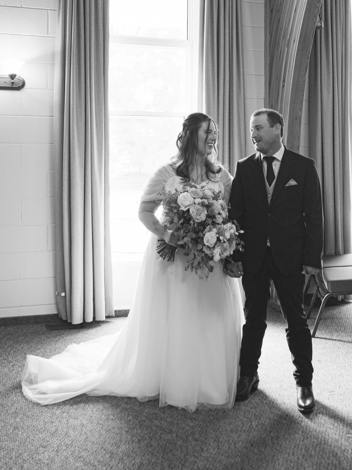 Black and white photo of a smiling bride in a white gown holding a bouquet beside a suited groom near a window.