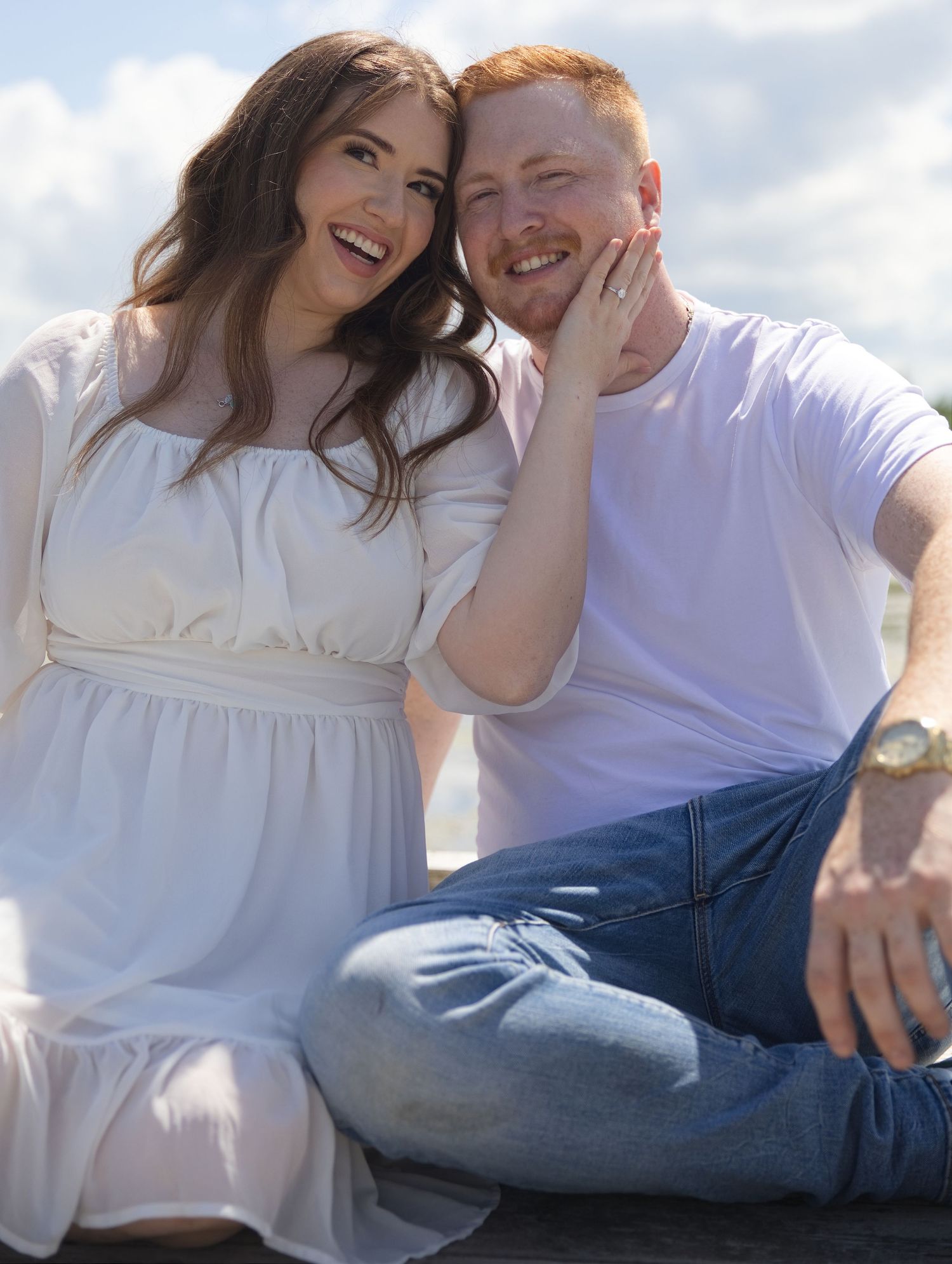 Happy couple sitting outdoors, woman in white dress and man in white t-shirt and jeans, smiling together on a sunny day.