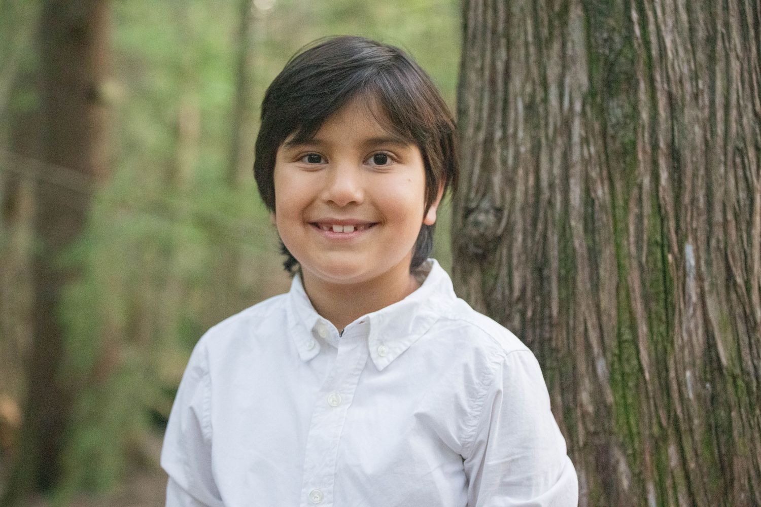 A smiling young boy with dark hair poses in front of a large tree.