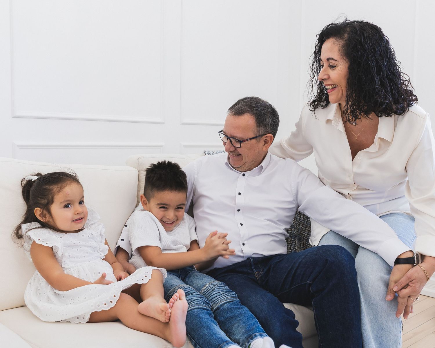 Happy family of four sitting on a white couch, parents smiling at each other while two young children sit beside them.