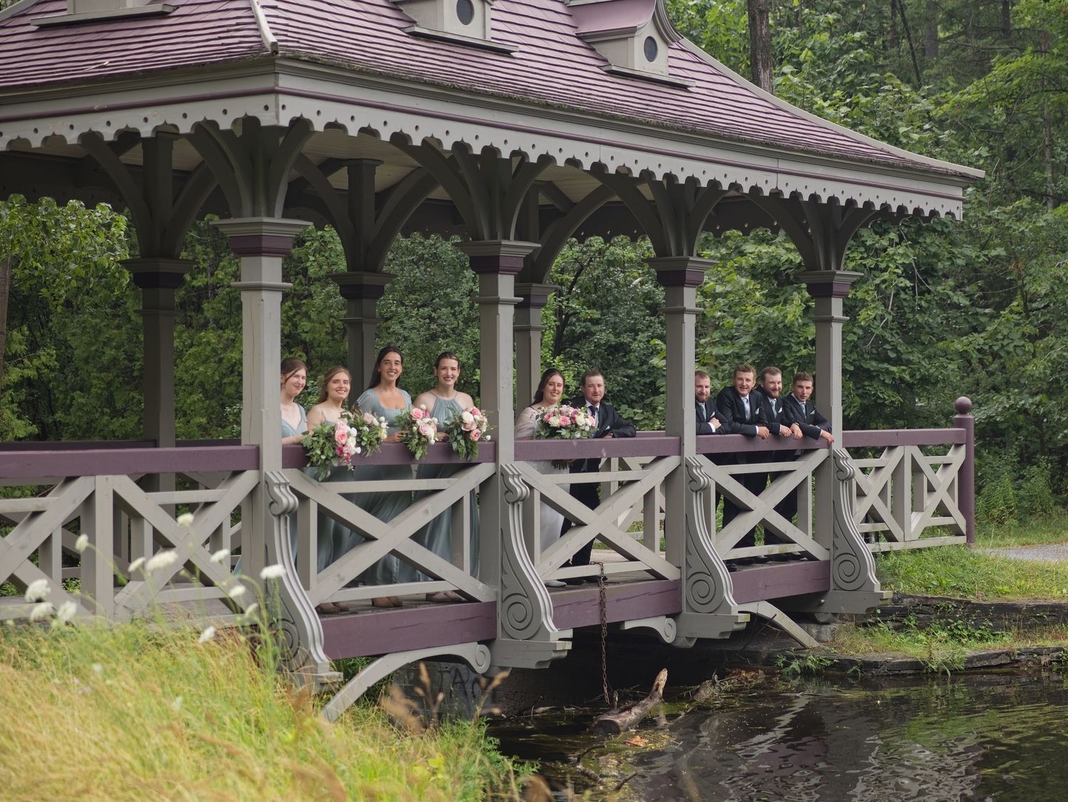 Wedding party poses on a charming purple gazebo bridge surrounded by lush green trees near a serene pond.