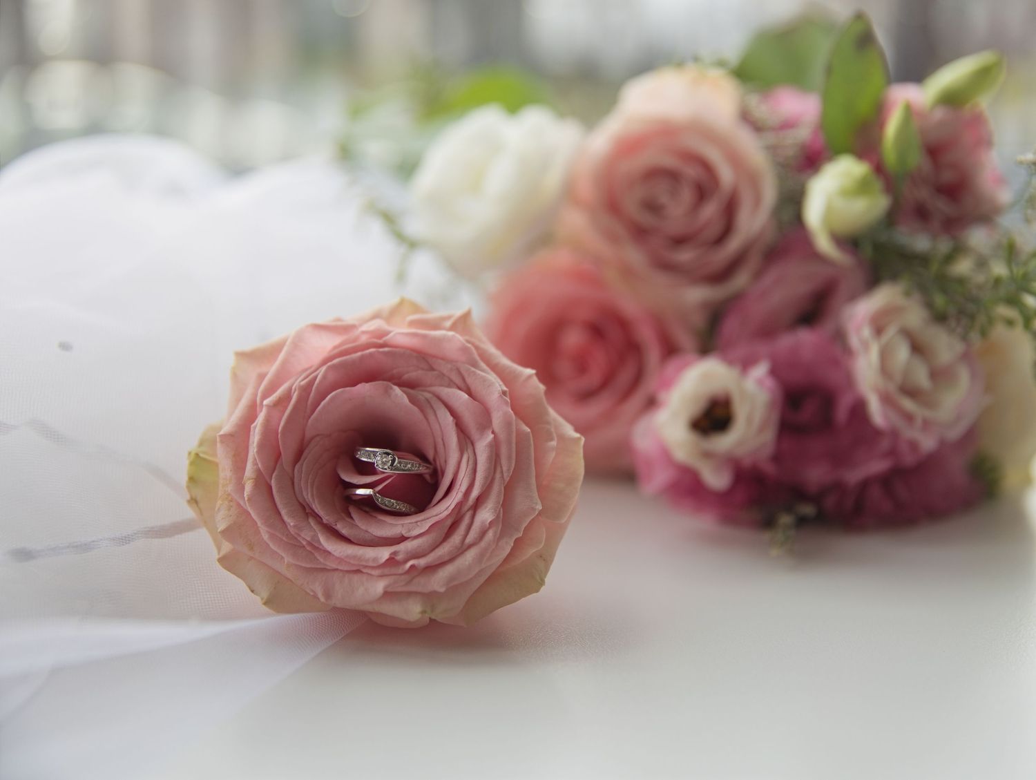 Diamond wedding rings nestled in a pink rose beside a romantic bridal bouquet of blush and white roses.