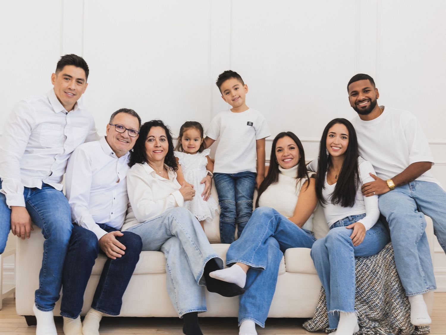 Happy extended family of eight posing together on a white couch in a bright studio.