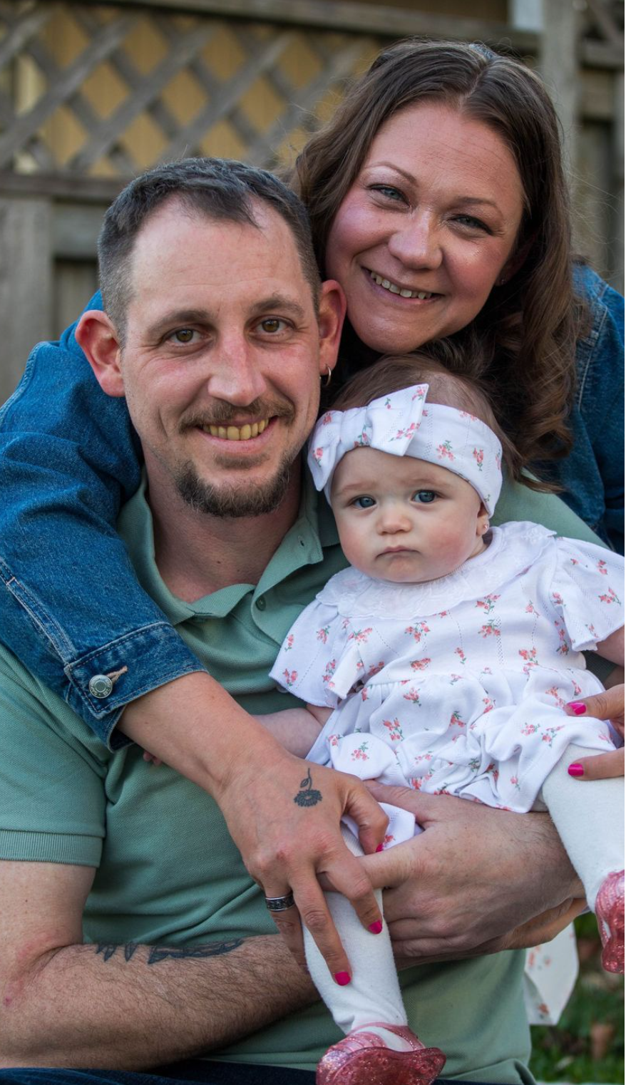 A smiling couple poses outdoors holding their baby girl in a white floral outfit with a bow headband.