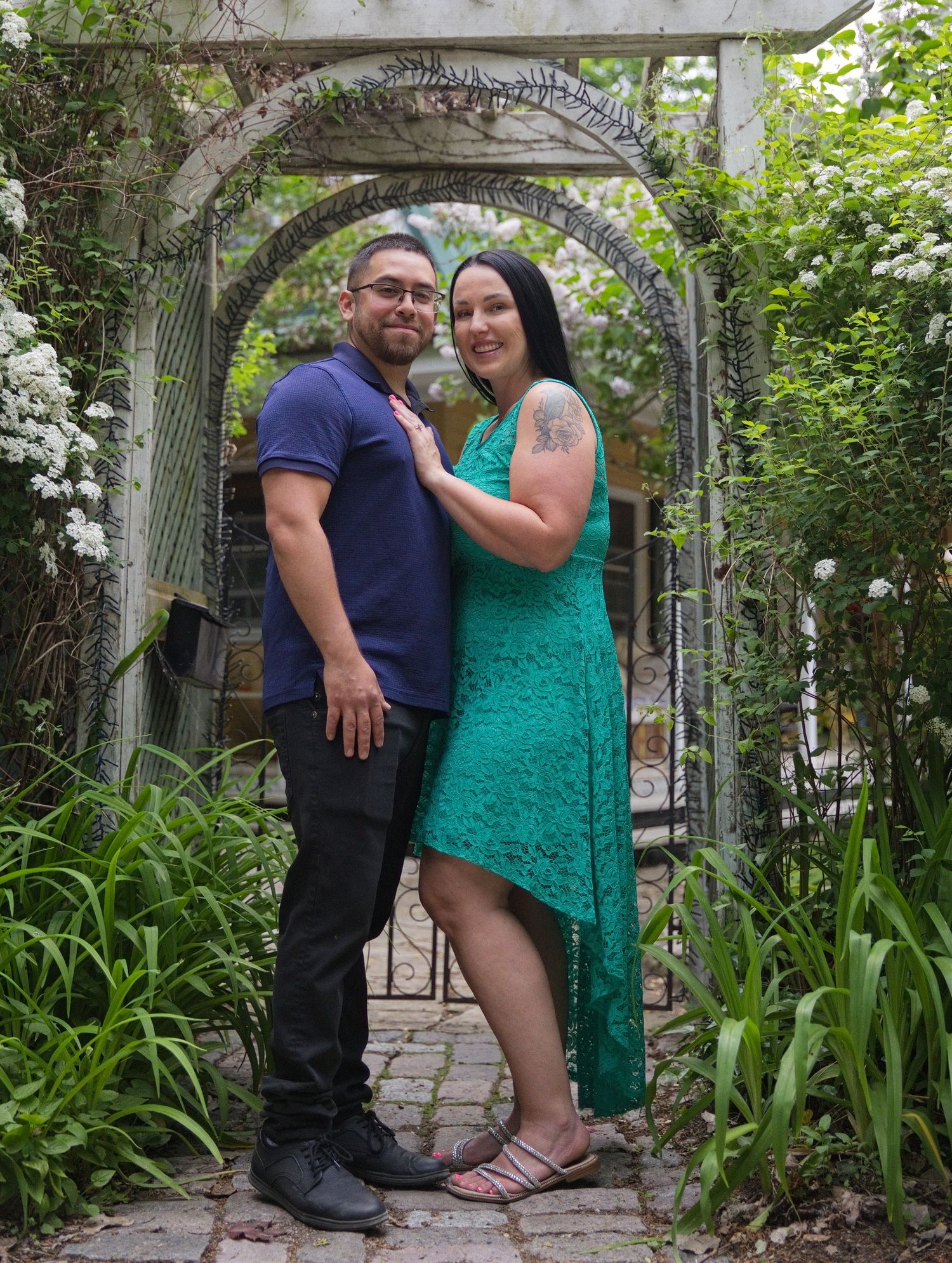 Couple posing under a garden archway, woman in teal lace dress and man in blue shirt, surrounded by lush greenery.