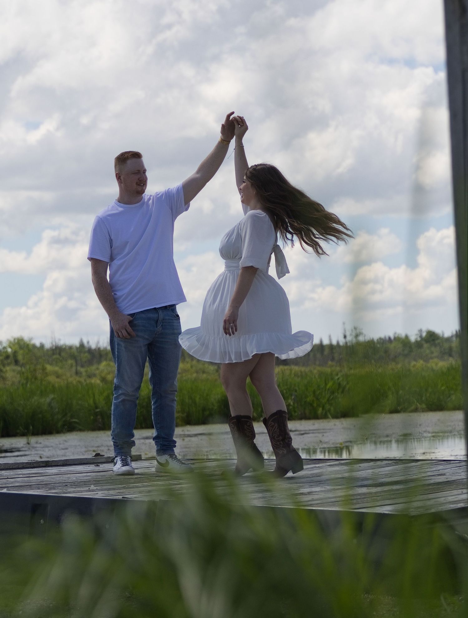 Couple dancing on a wooden dock surrounded by lush greenery and cloudy skies, woman in white dress and boots.