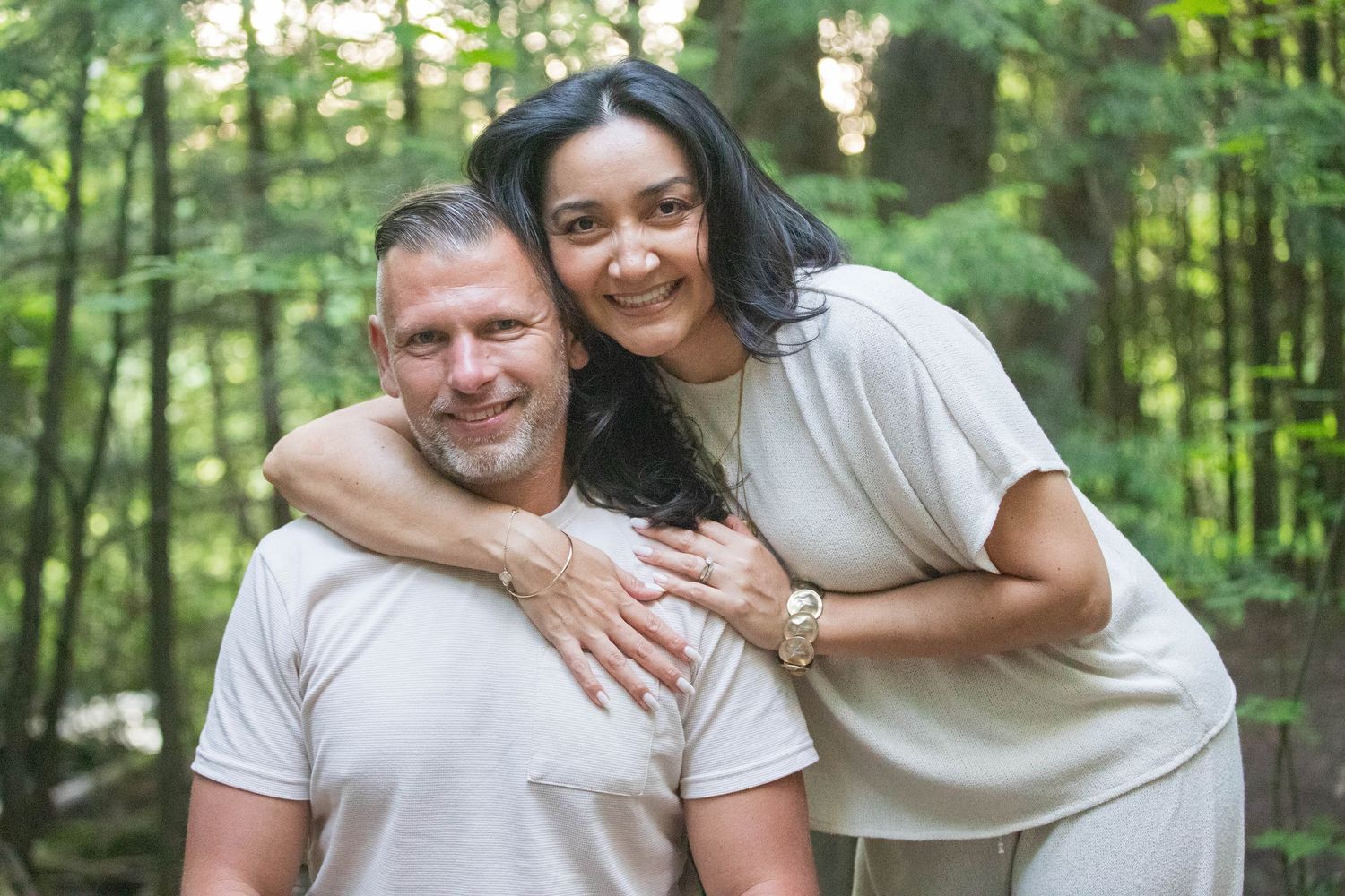 A smiling couple embraces warmly in a lush green forest setting.