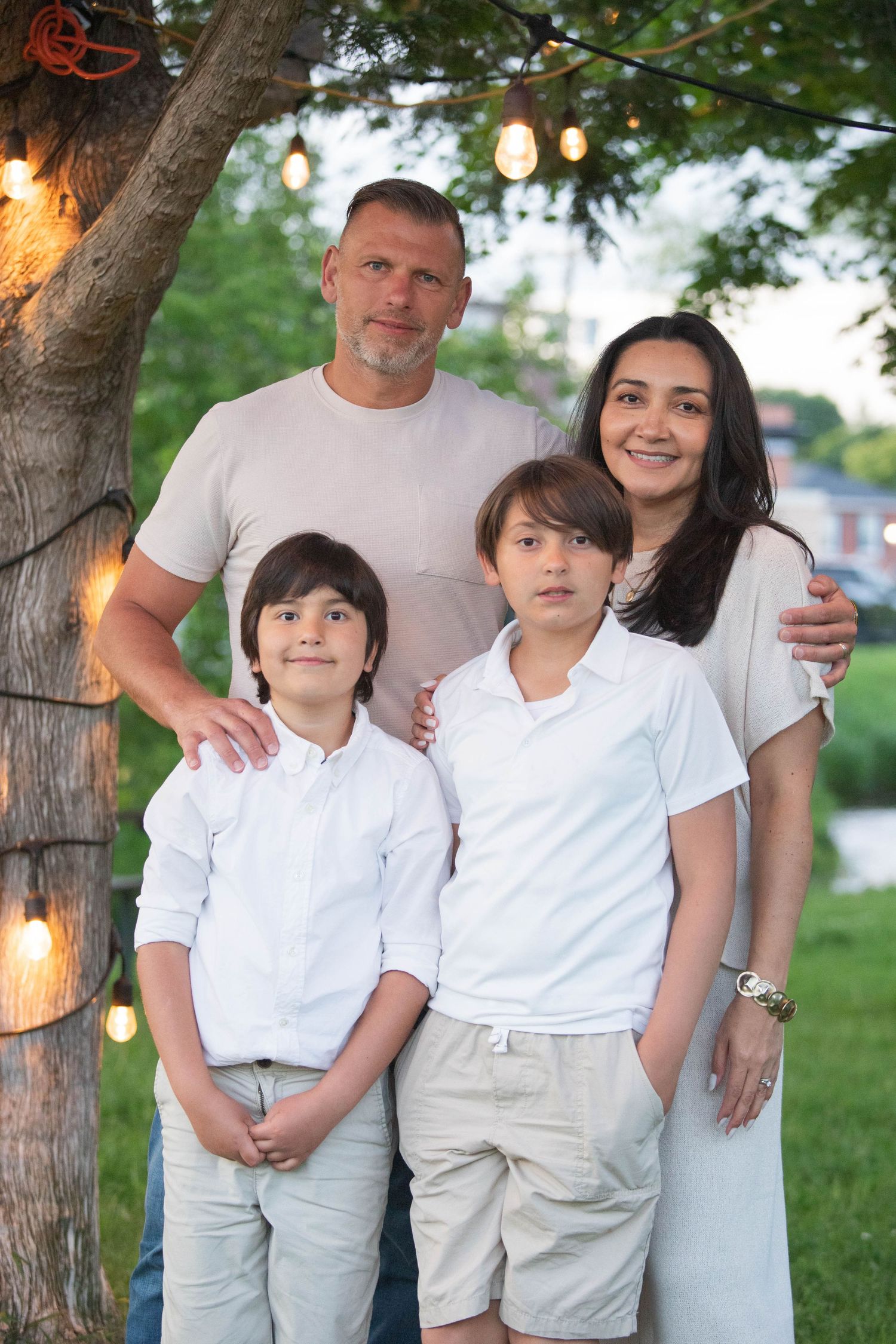 Family of four posing under string lights by a tree in a lush green outdoor setting.