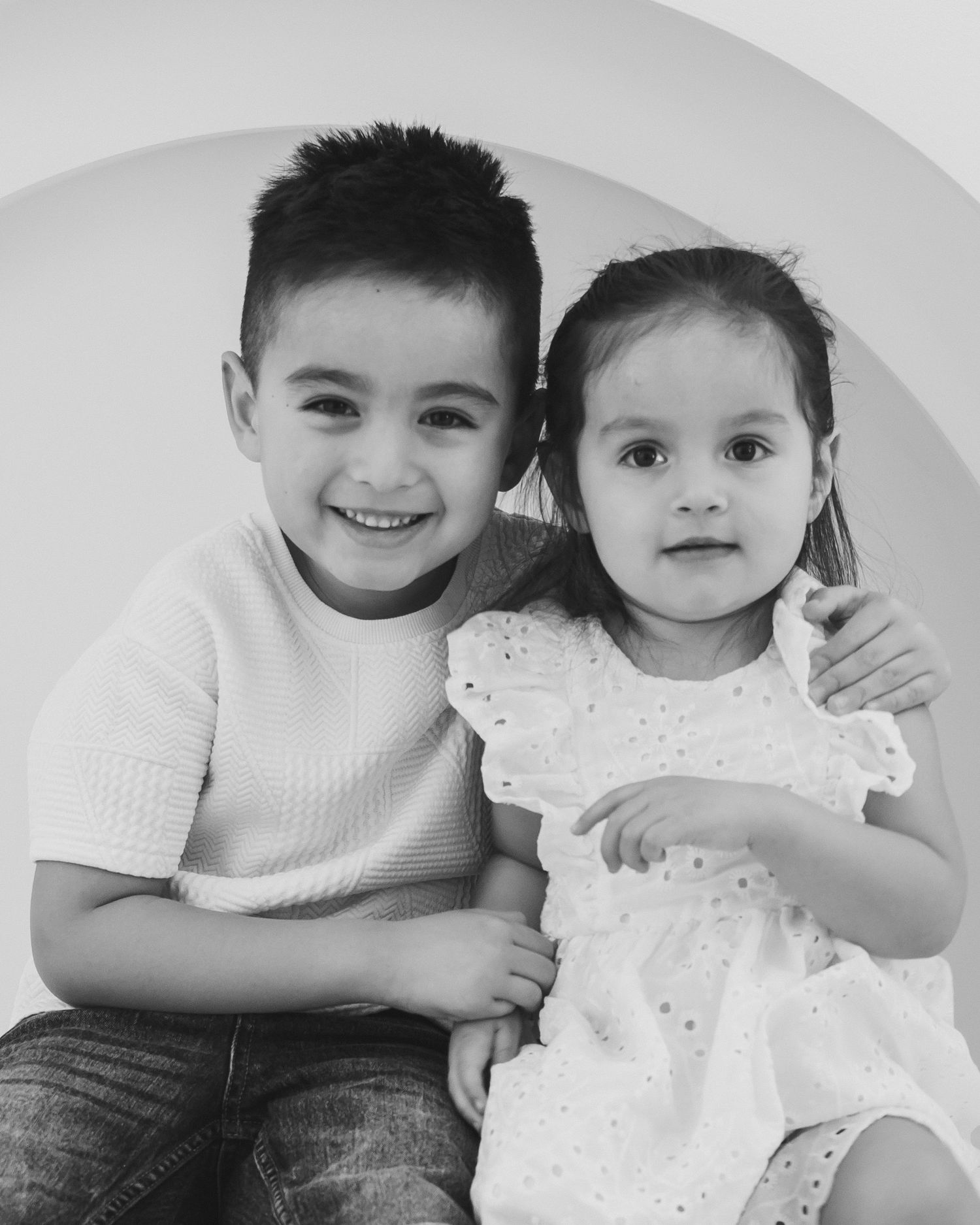 Black and white portrait of two young siblings, boy and toddler girl, posing together.