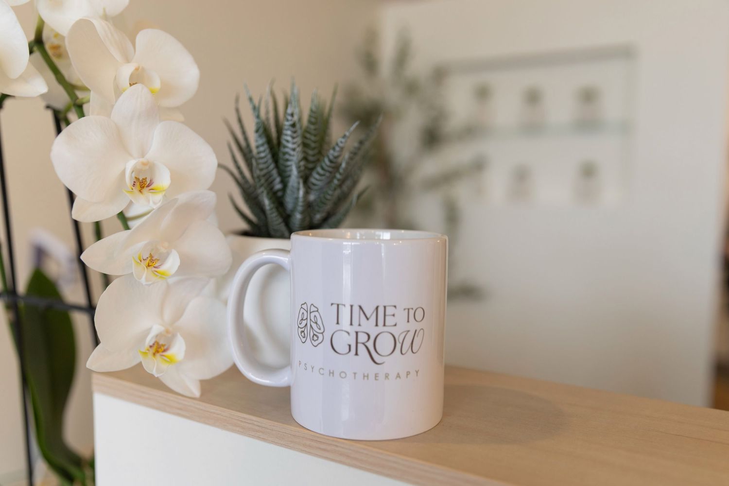 White mug with Time to Grow logo on wooden shelf beside white orchids and succulent plant.