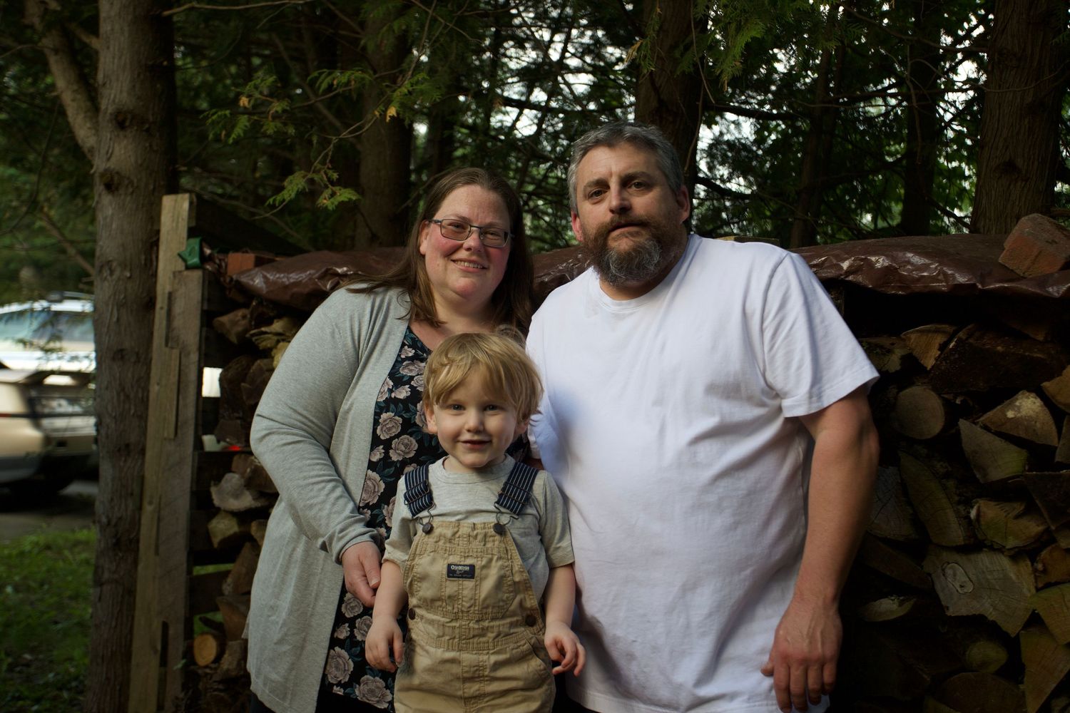 Family of three posing together in front of stacked firewood outdoors.