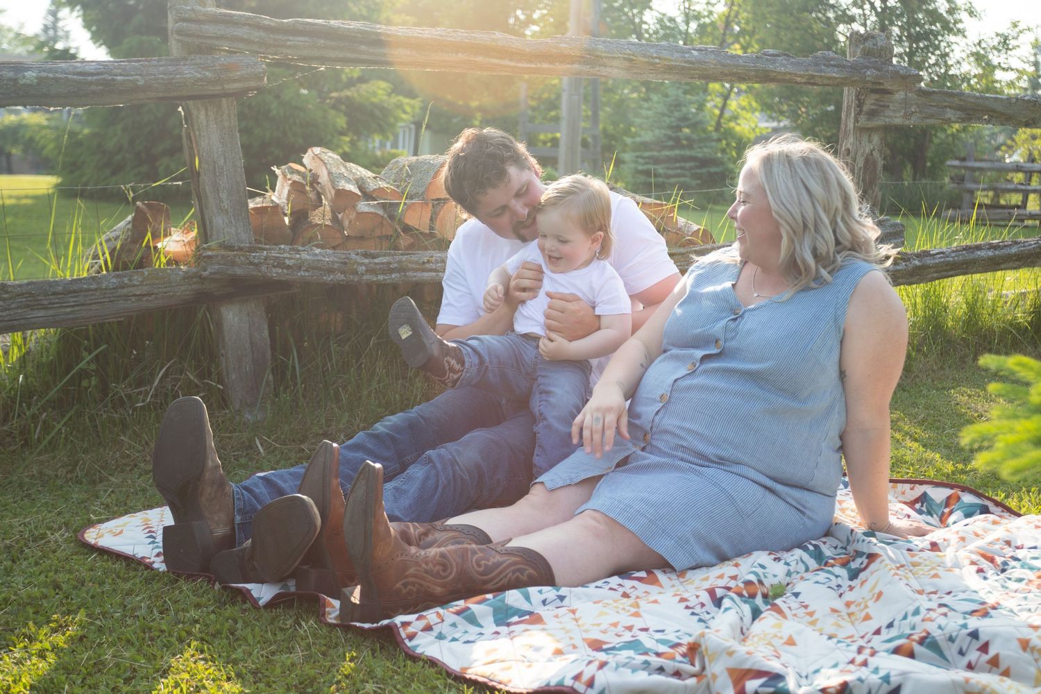A couple relaxes on a colorful blanket outdoors in golden sunlight while holding their baby.