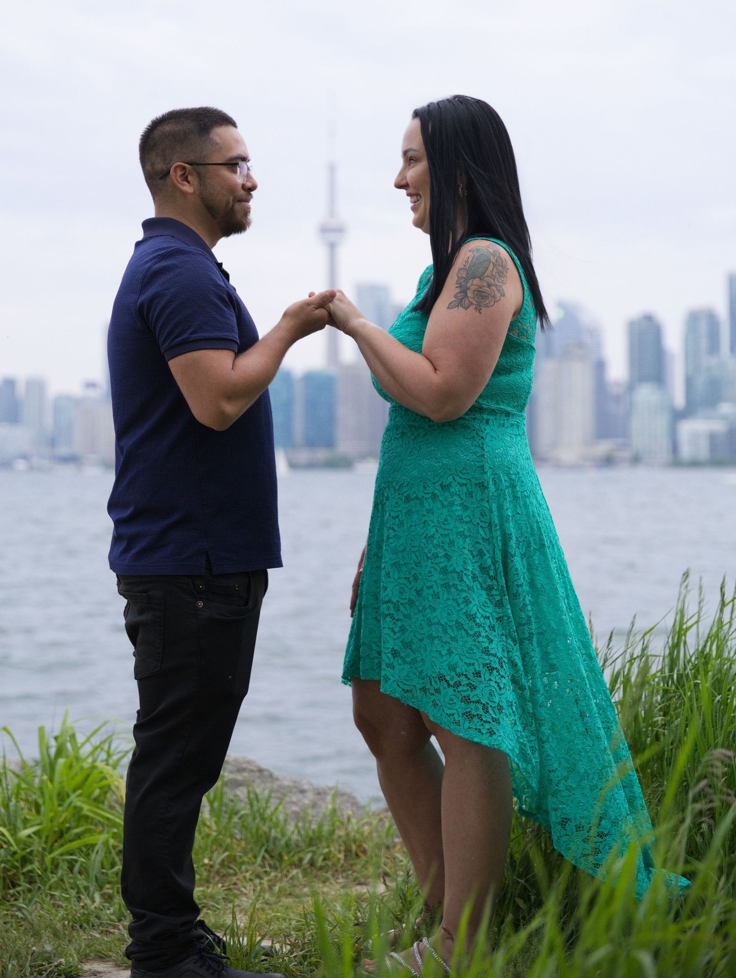 Couple holding hands by Toronto waterfront with CN Tower skyline backdrop, man in navy shirt and woman in teal lace dress.