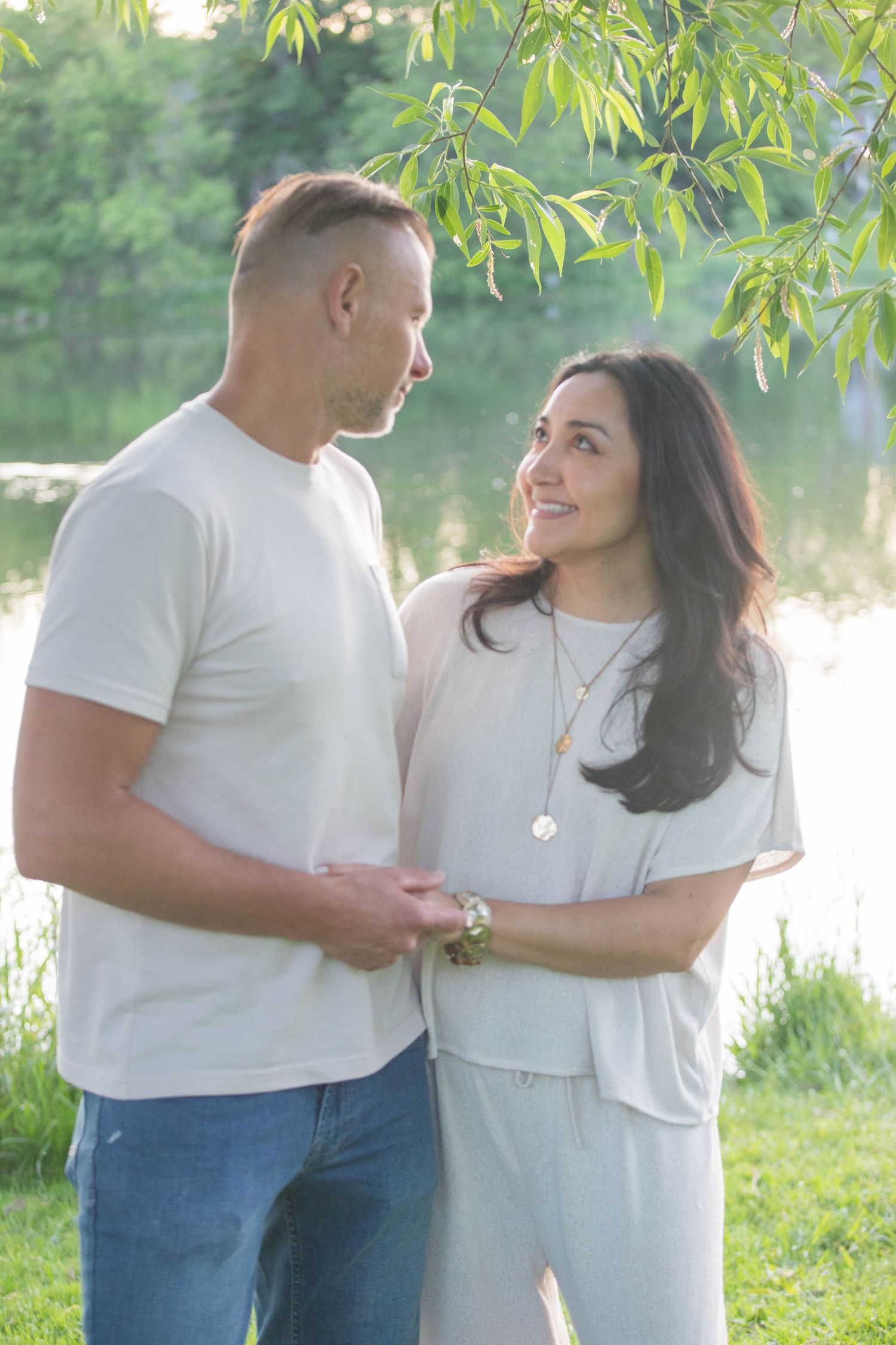 Romantic couple holding hands and gazing at each other near a serene lake under willow tree branches.
